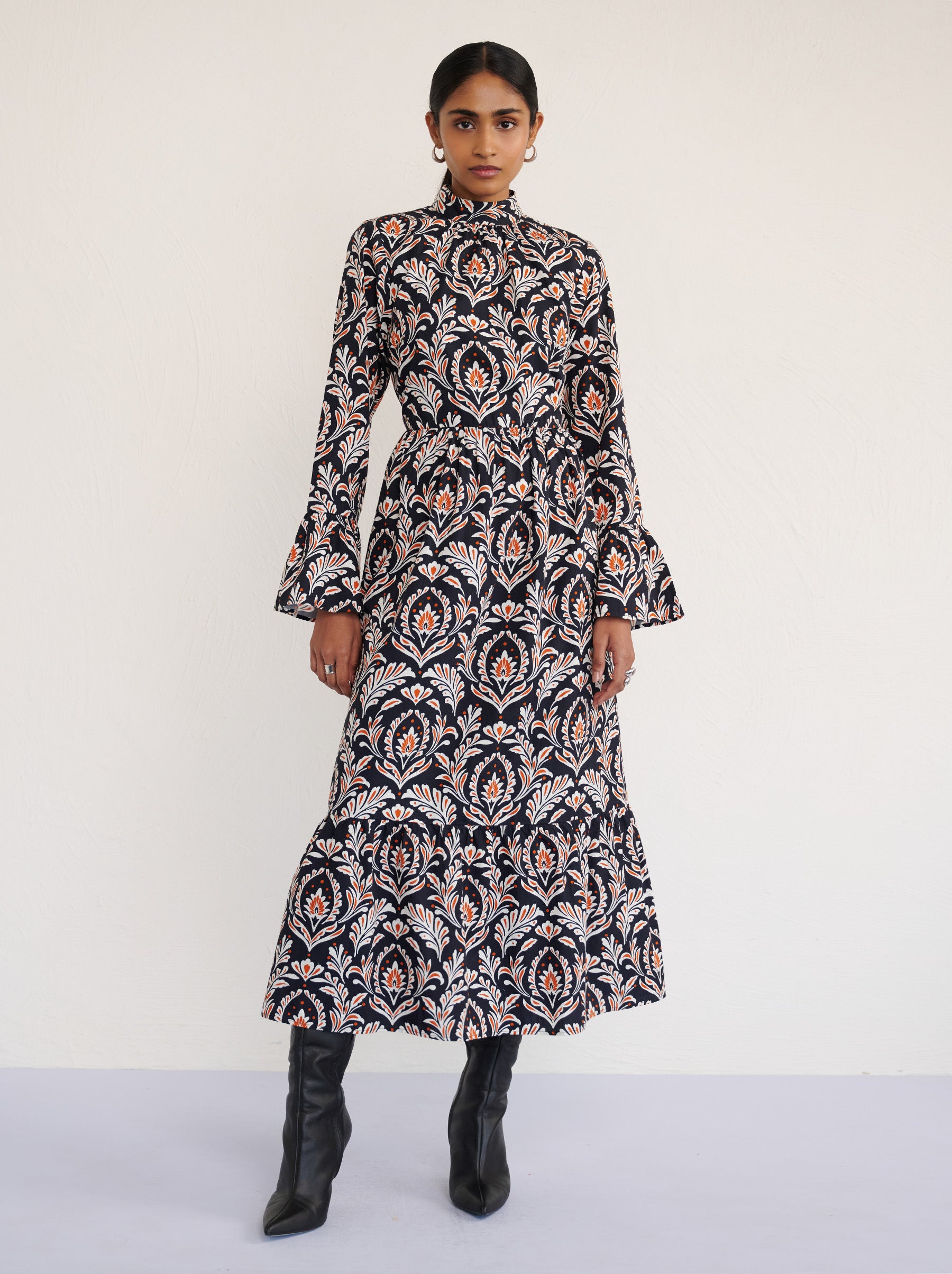 Front view of a model standing. She is wearing the Sumi long-sleeve dress in crisp cotton poplin in Akbar Galaxy print. It is featuring a high neckline, ruffled sleeves, a flowing skirt, and side-seam pockets.