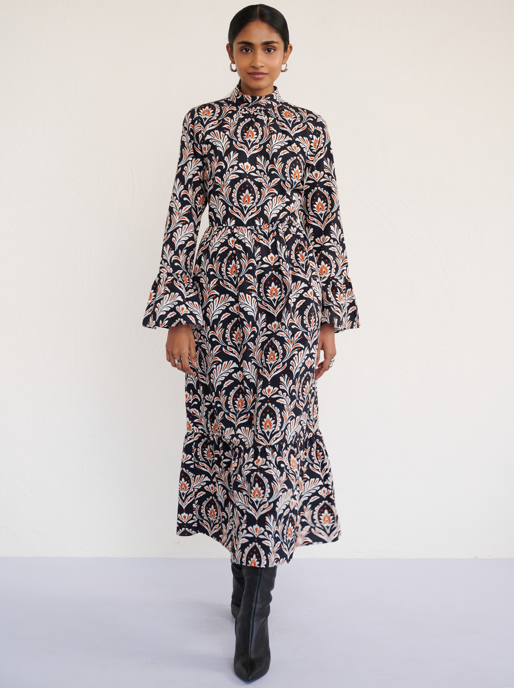 Front view of a model walking. She is wearing the Sumi long-sleeve dress in crisp cotton poplin in Akbar Galaxy print. It is featuring a high neckline, ruffled sleeves, a flowing skirt, and side-seam pockets.