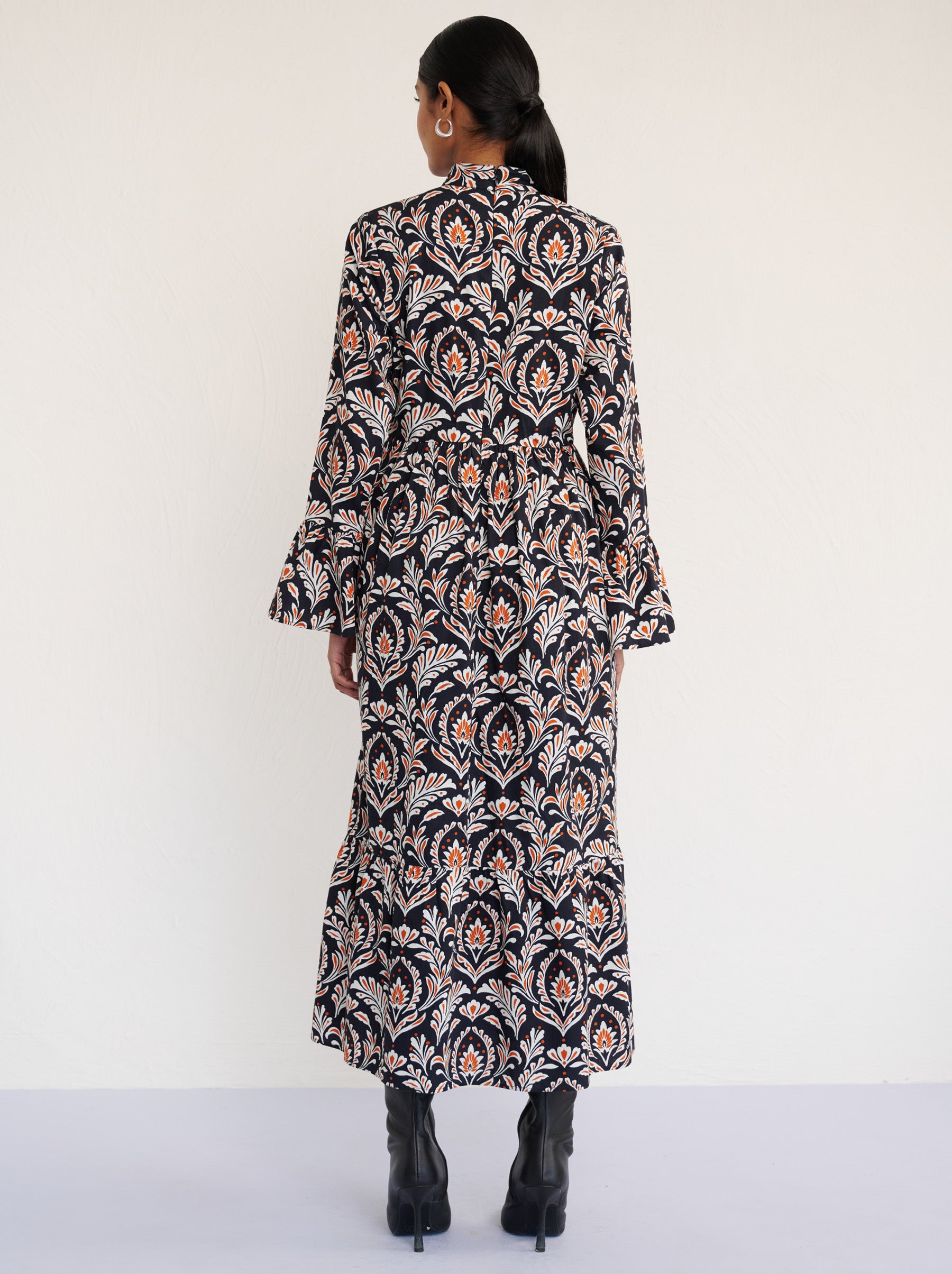 Back view of a model standing. She is wearing the Sumi long-sleeve dress in crisp cotton poplin in Akbar Galaxy print. It is featuring a high neckline, ruffled sleeves, a flowing skirt, and side-seam pockets.