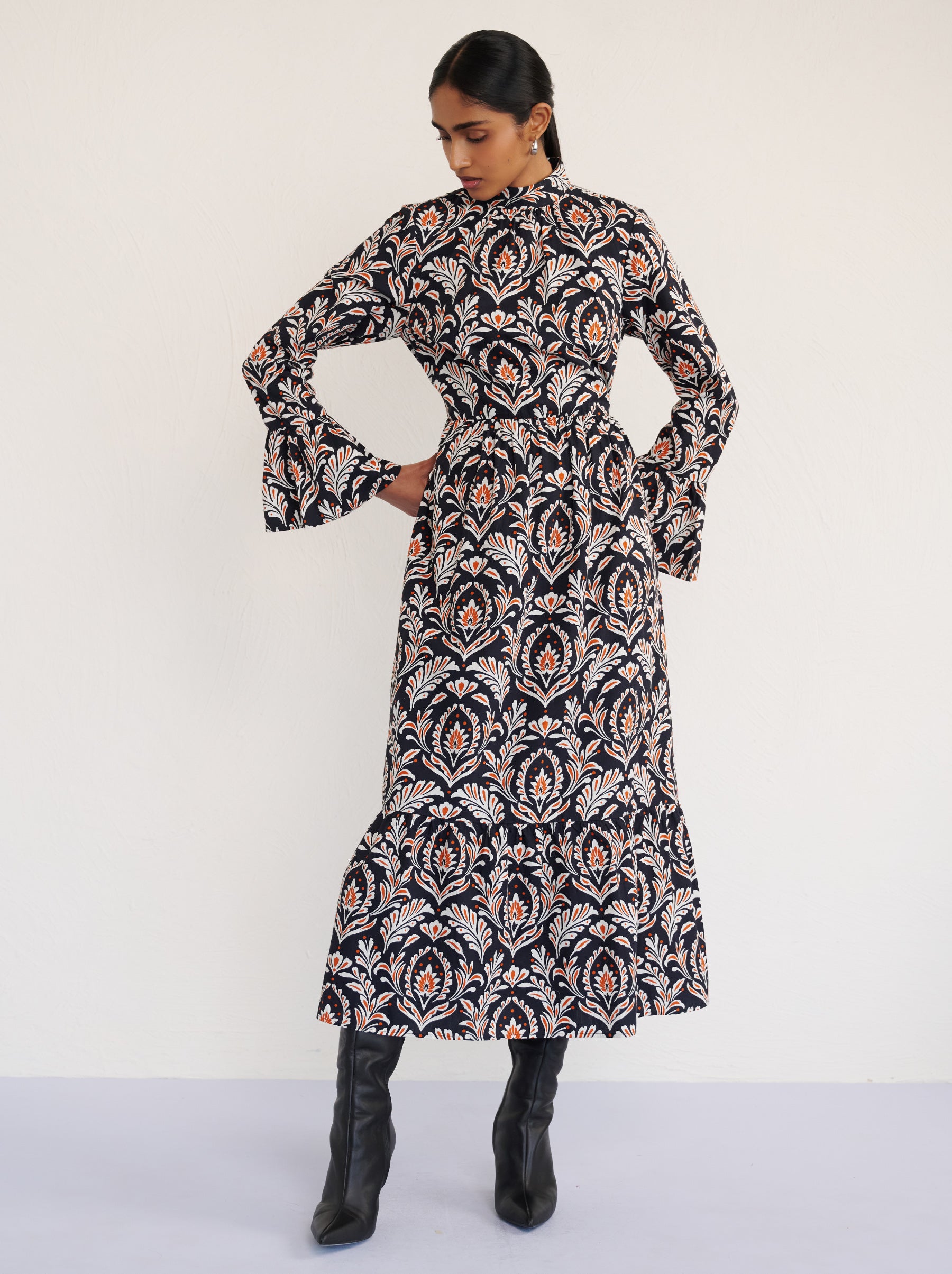 Front view of a model standing. She is wearing the Sumi long-sleeve dress in crisp cotton poplin in Akbar Galaxy print. It is featuring a high neckline, ruffled sleeves, a flowing skirt, and side-seam pockets.