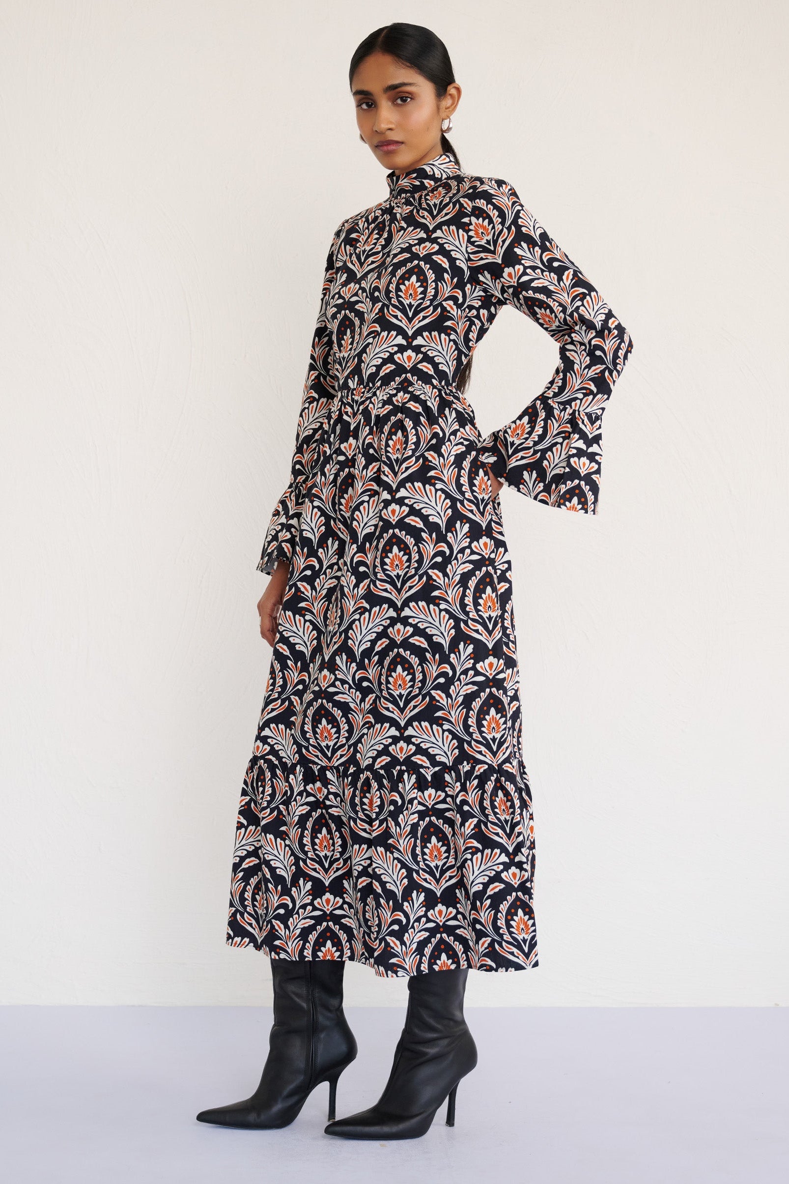 Side view of a model standing with right hand in pocket. She is wearing the Sumi long-sleeve dress in crisp cotton poplin in Akbar Galaxy print. It is featuring a high neckline, ruffled sleeves, a flowing skirt, and side-seam pockets.