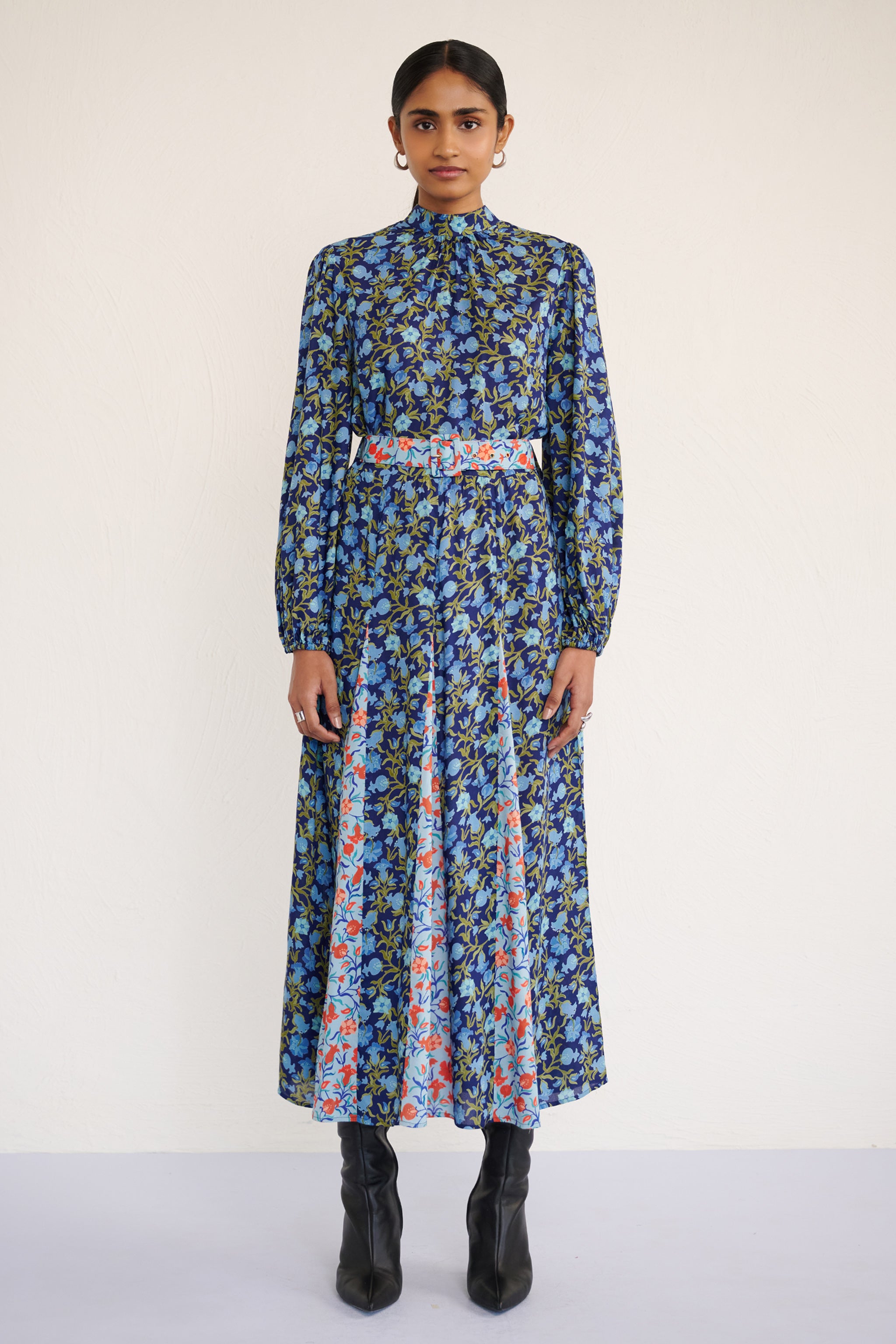 Front view of woman wearing a full-length dress with a stand collar, fitted waist belt, and flowing skirt featuring contrast print inserts and a pomegranate vine motif.