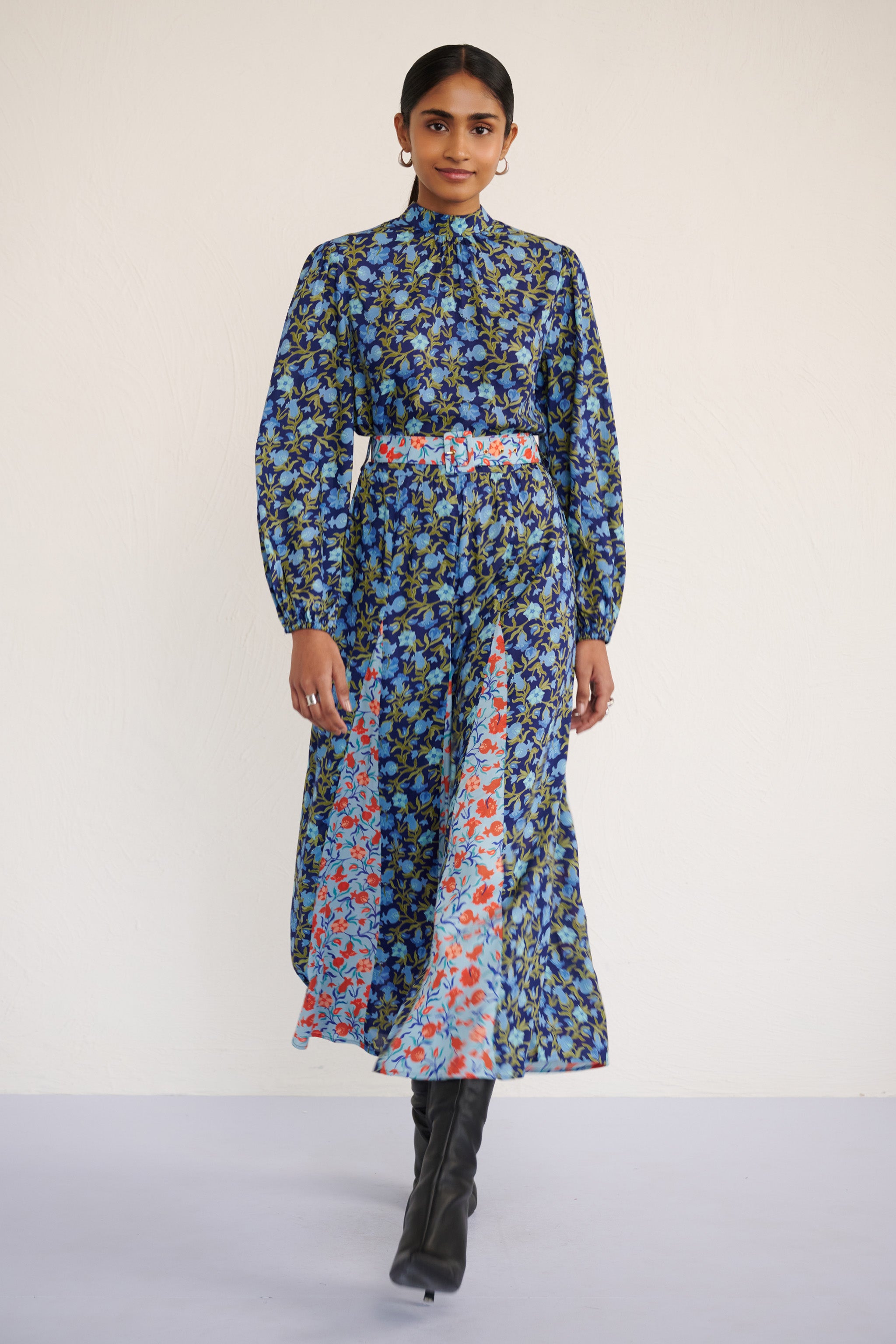 Front view of woman wearing a full-length dress with a stand collar, fitted waist belt, and flowing skirt featuring contrast print inserts and a pomegranate vine motif.
