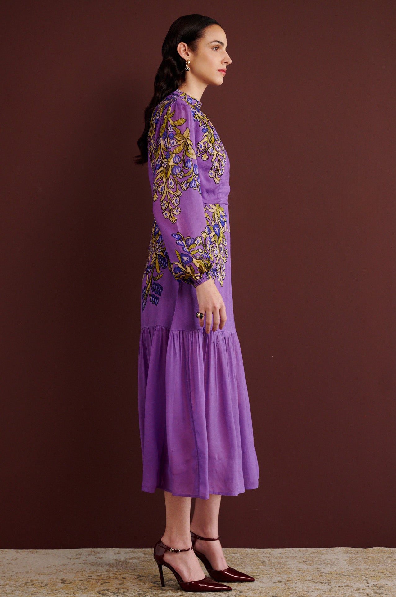 Side view of woman standing. She is wearing the Sumina dress in the Unicorn Tapestry print. It is a purple viscose voile midi dress with a fitted crew neck, and tiered skirt, featuring intricate floral and mythical motifs inspired by 15th-century Scottish embroidery.