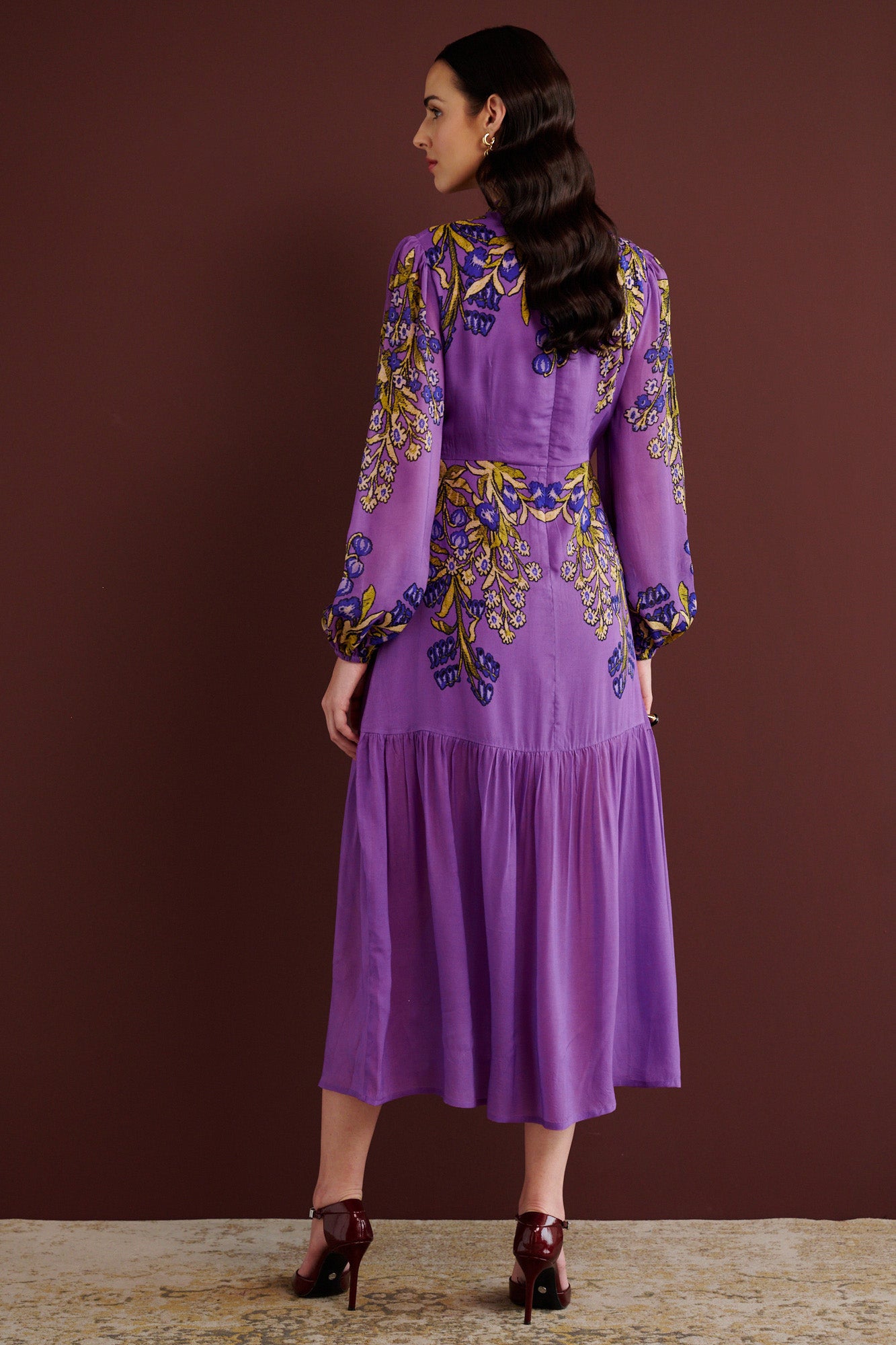 Back view of woman standing. She is wearing the Sumina dress in the Unicorn Tapestry print. It is a purple viscose voile midi dress with a fitted crew neck, and tiered skirt, featuring intricate floral and mythical motifs inspired by 15th-century Scottish embroidery.