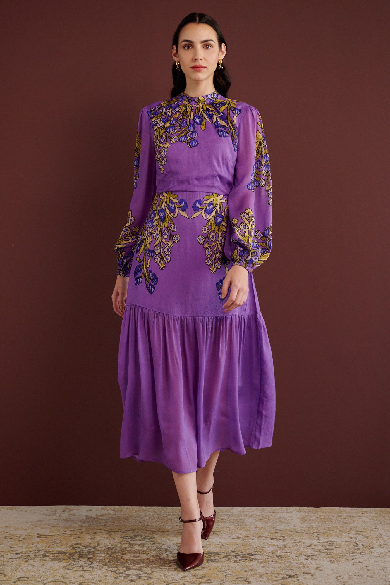 Front view of woman walking. She is wearing the Sumina dress in the Unicorn Tapestry print. It is a purple viscose voile midi dress with a fitted crew neck, and tiered skirt, featuring intricate floral and mythical motifs inspired by 15th-century Scottish embroidery.