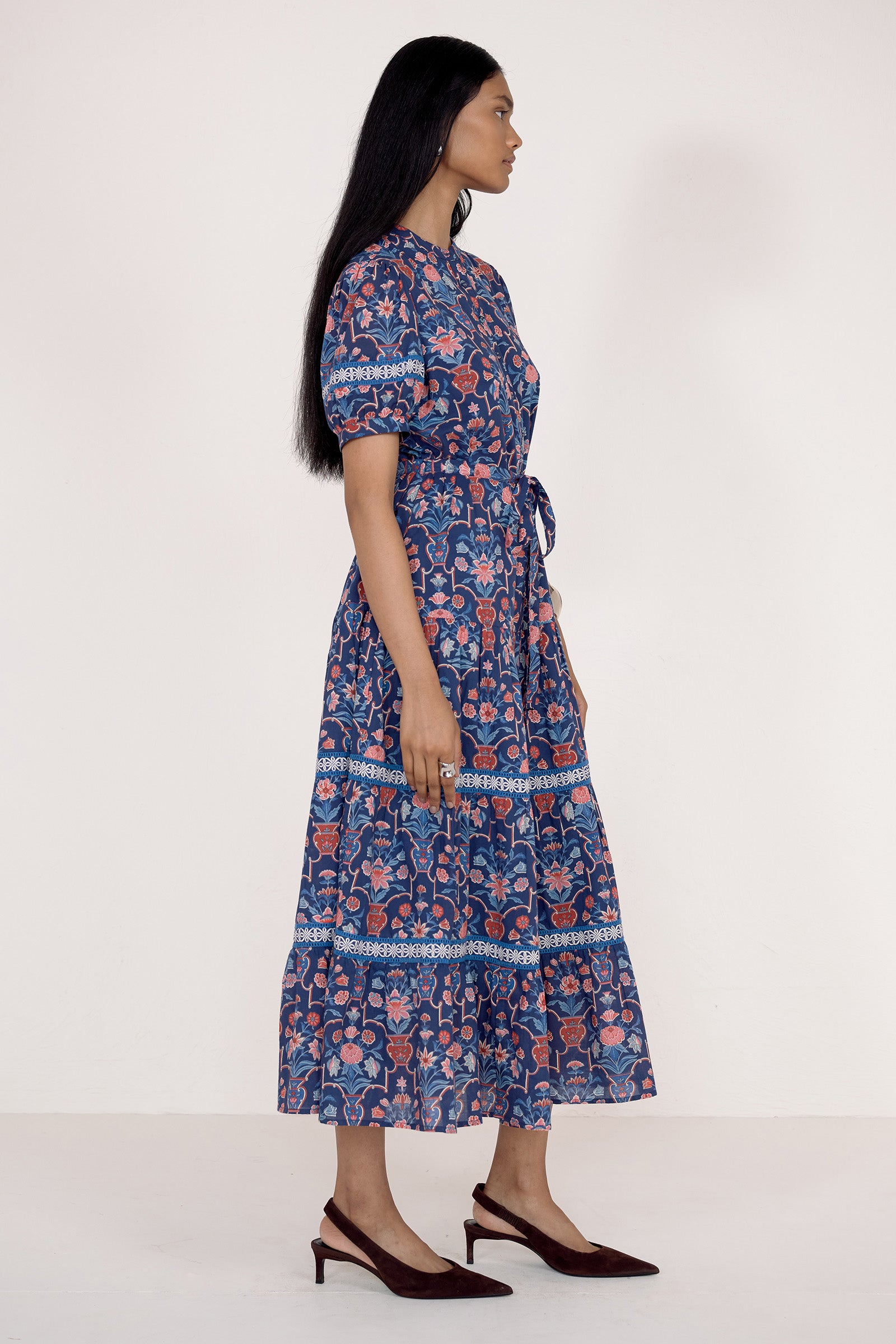 Side view of model standing wearing the Saylor dress in Floral Vase print. It features a shirtdress silhouette, lace trim, tiered skirt, and detachable waist belt.