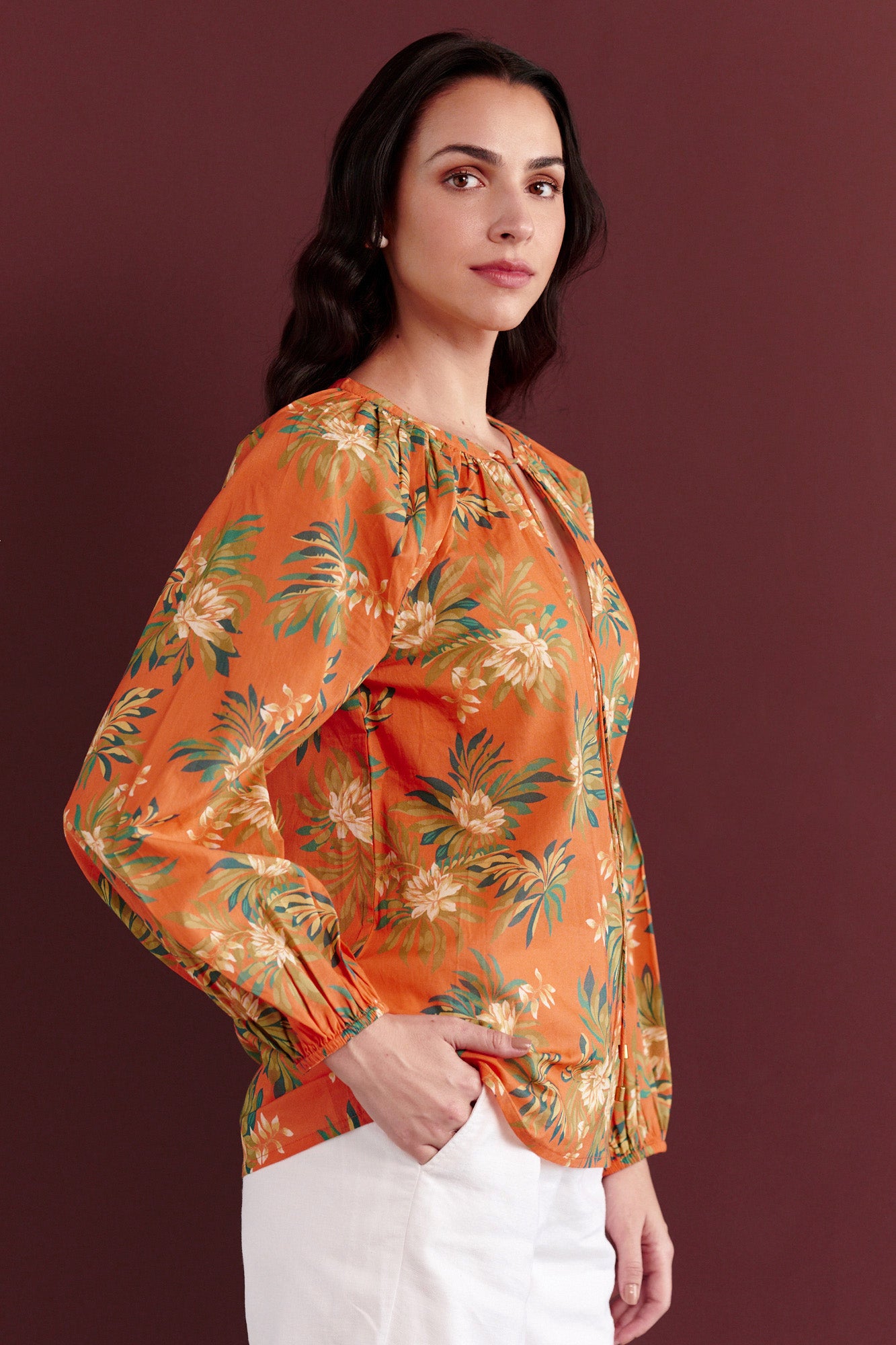 Side view of woman standing. She is wearing the Tara crew-neck shirt in the Artichoke Flower print paired with white trouser. It is a breezy cotton voile top with a tie-neck detail and a relaxed, tropical-inspired design.
