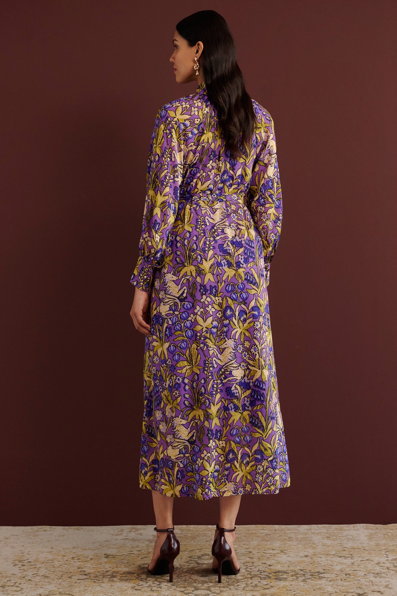 Back view of woman standing. She is wearing the Tara dress in the Unicorn Tapestry print. It is a flowing purple viscose voile dress with a stand collar, gathered neckline and sleeves, fabric-covered buttons, and a removable belt, featuring intricate floral and mythical motifs inspired by the Scottish Highlands.