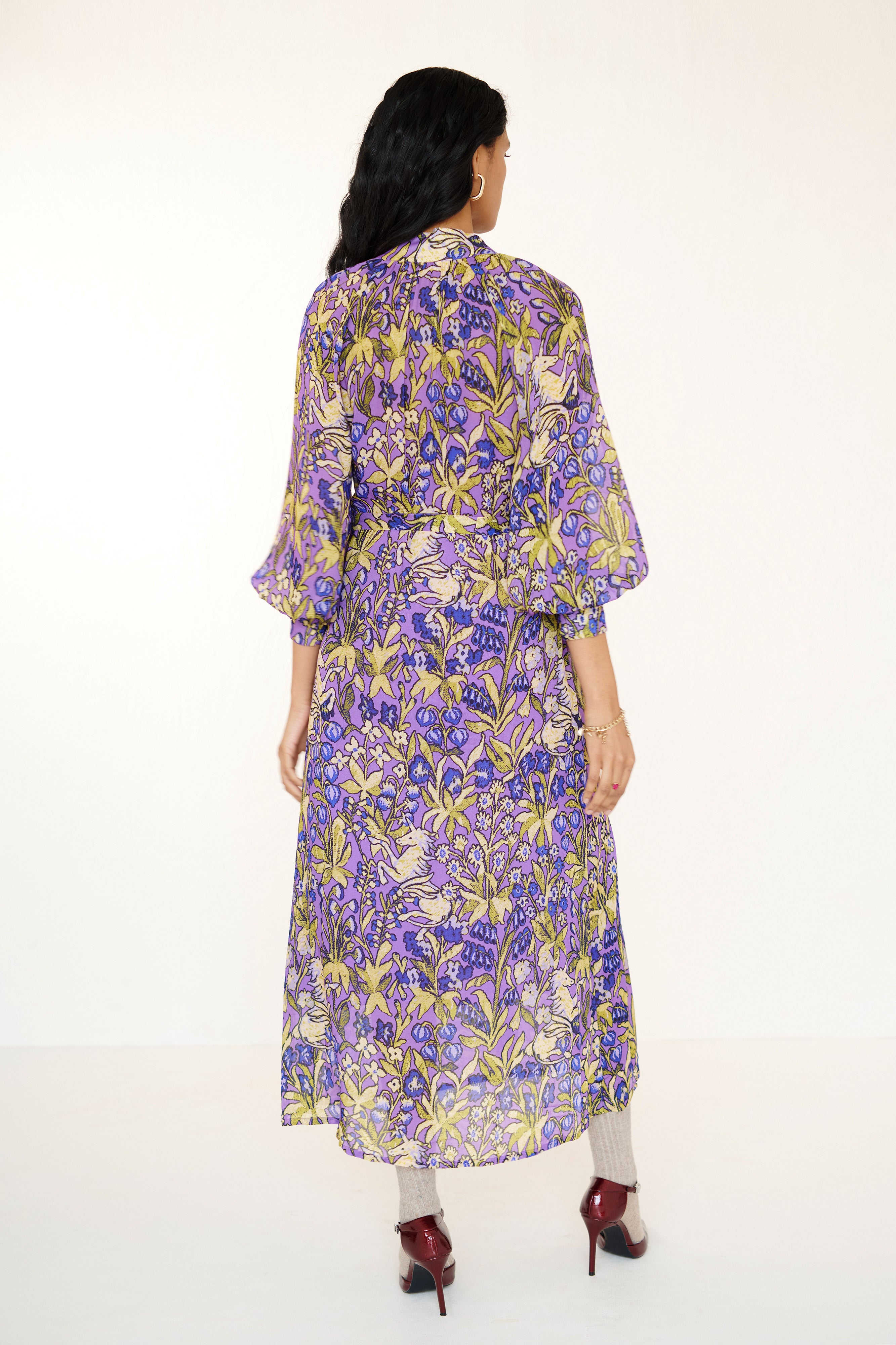 Back view of woman standing. She is wearing the Tara dress in the Unicorn Tapestry print. It is a flowing purple viscose voile dress with a stand collar, gathered neckline and sleeves, fabric-covered buttons, and a removable belt, featuring intricate floral and mythical motifs inspired by the Scottish Highlands.
