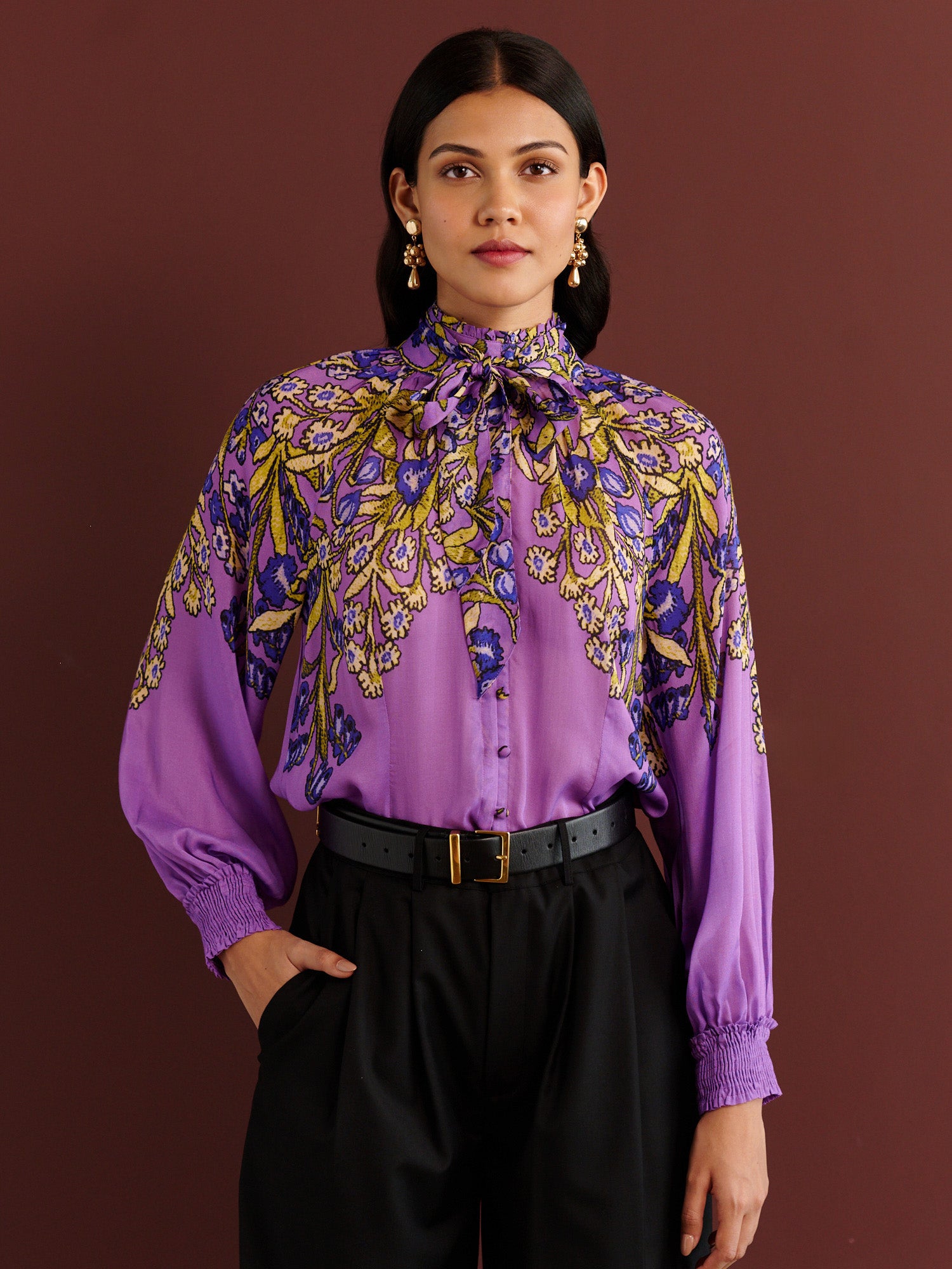 Front view of woman standing. She is wearing the Tara tie-neck shirt in the Unicorn Tapestry print, a purple viscose voile blouse with yellow and blue floral motifs, high ruffled collar, and gathered sleeves, paired with black pleated trousers and a black belt.