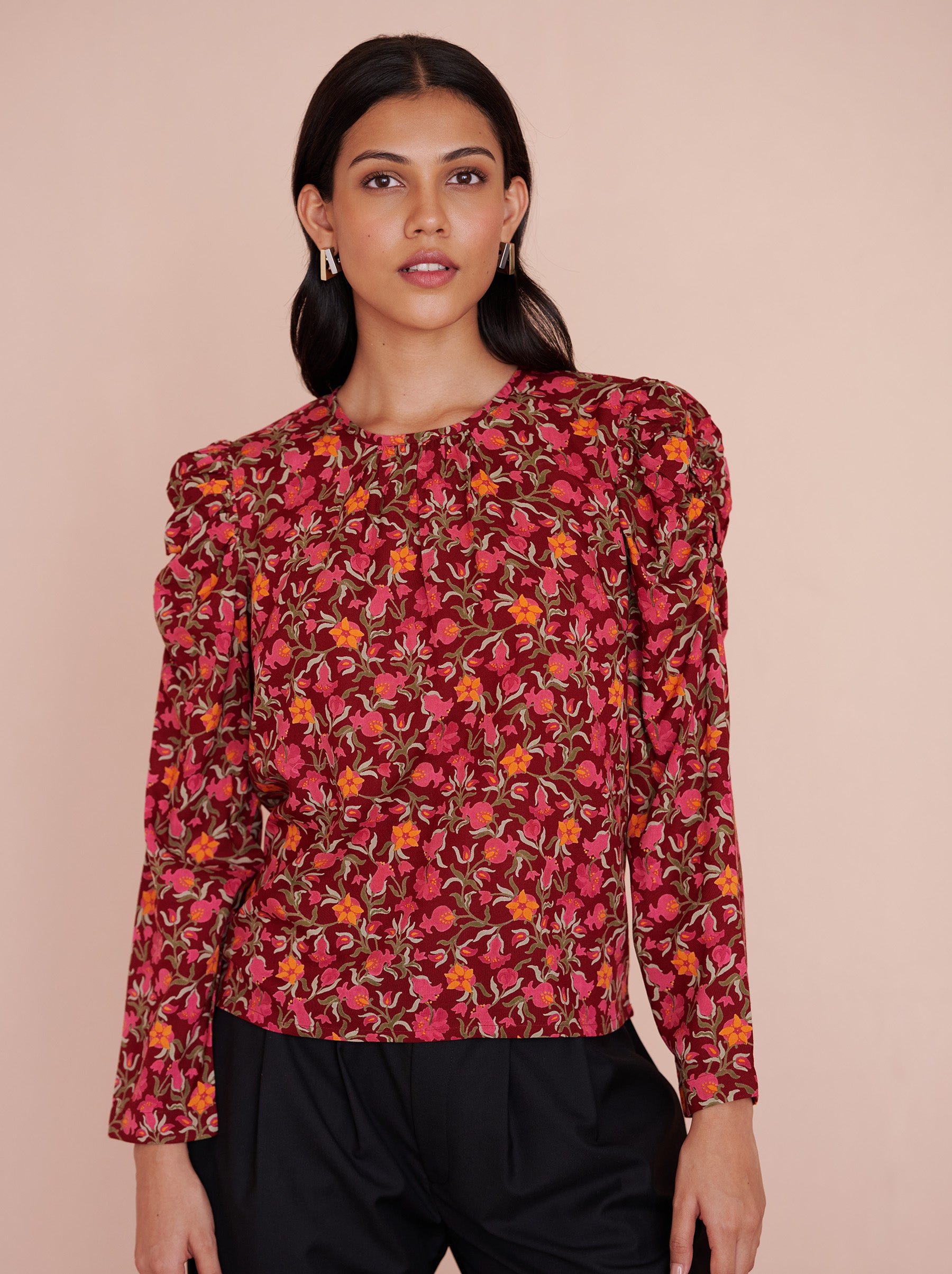 Front view close up of a model standing. She is wearing the Vincent top in eco-friendly viscose, featuring the Pomegranate Vine print in Azalea color. It has soft gathers at the neck and shoulders, long sleeves, and a crew neckline. The elegant day-to-night style fastens with a back zipper.