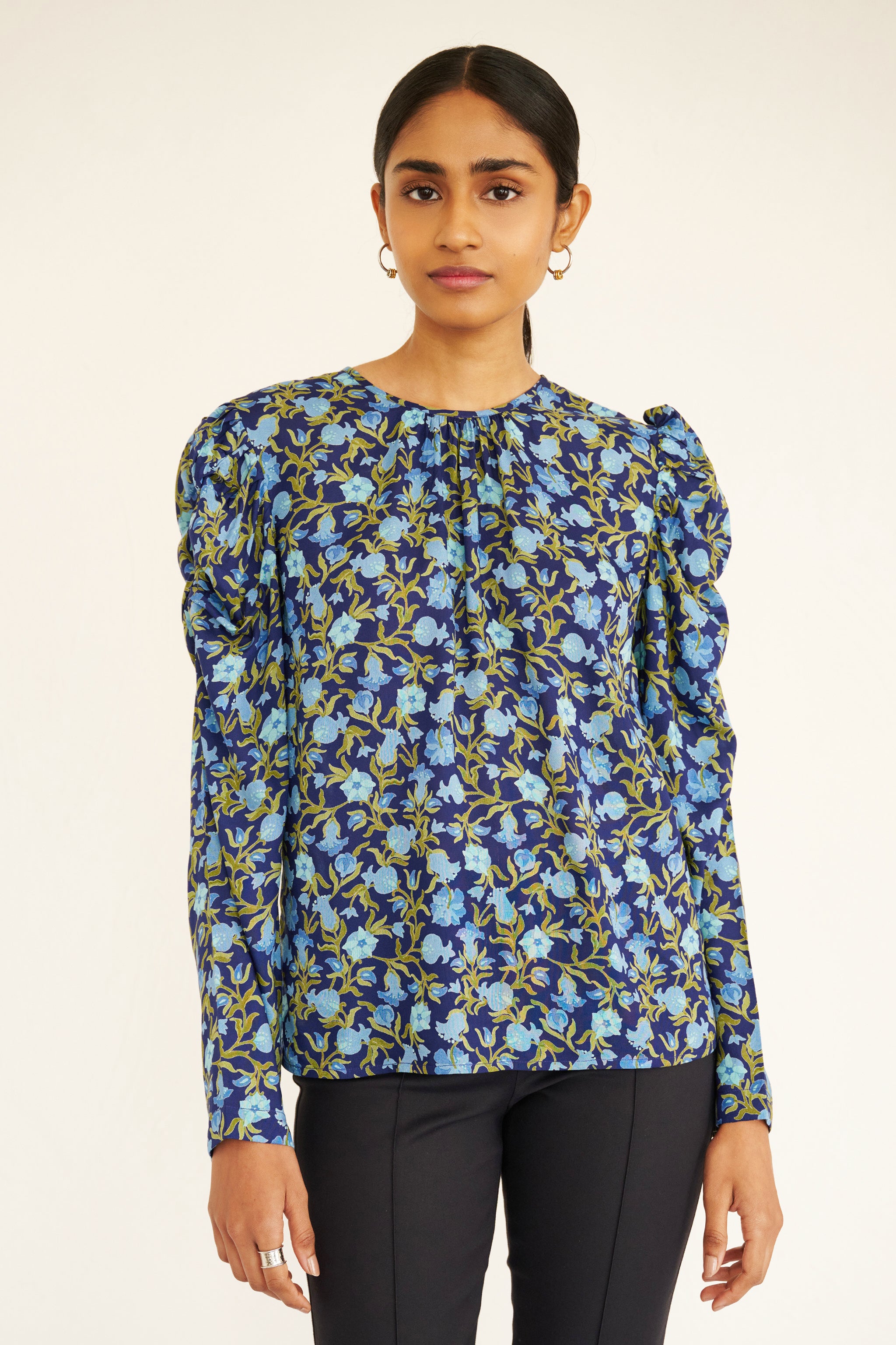 Front view of woman wearing a navy blue blouse with light blue and green pomegranate vine print, featuring soft gathers at the neck and puffed shoulders, paired with slim black trousers and gold hoop earrings.