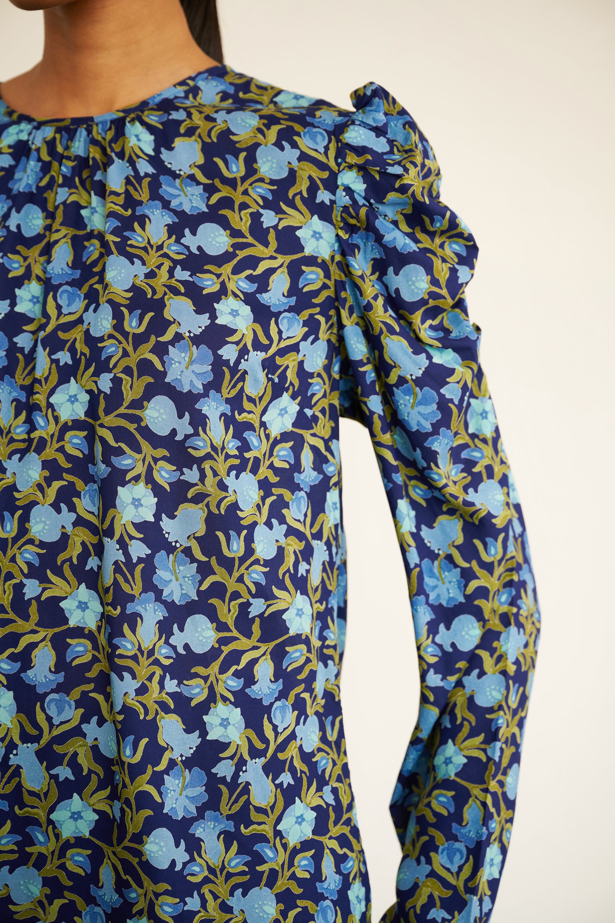 Sleeve detail close up of woman wearing a navy blue blouse with light blue and green pomegranate vine print, featuring soft gathers at the neck and puffed shoulders, paired with slim black trousers and gold hoop earrings.