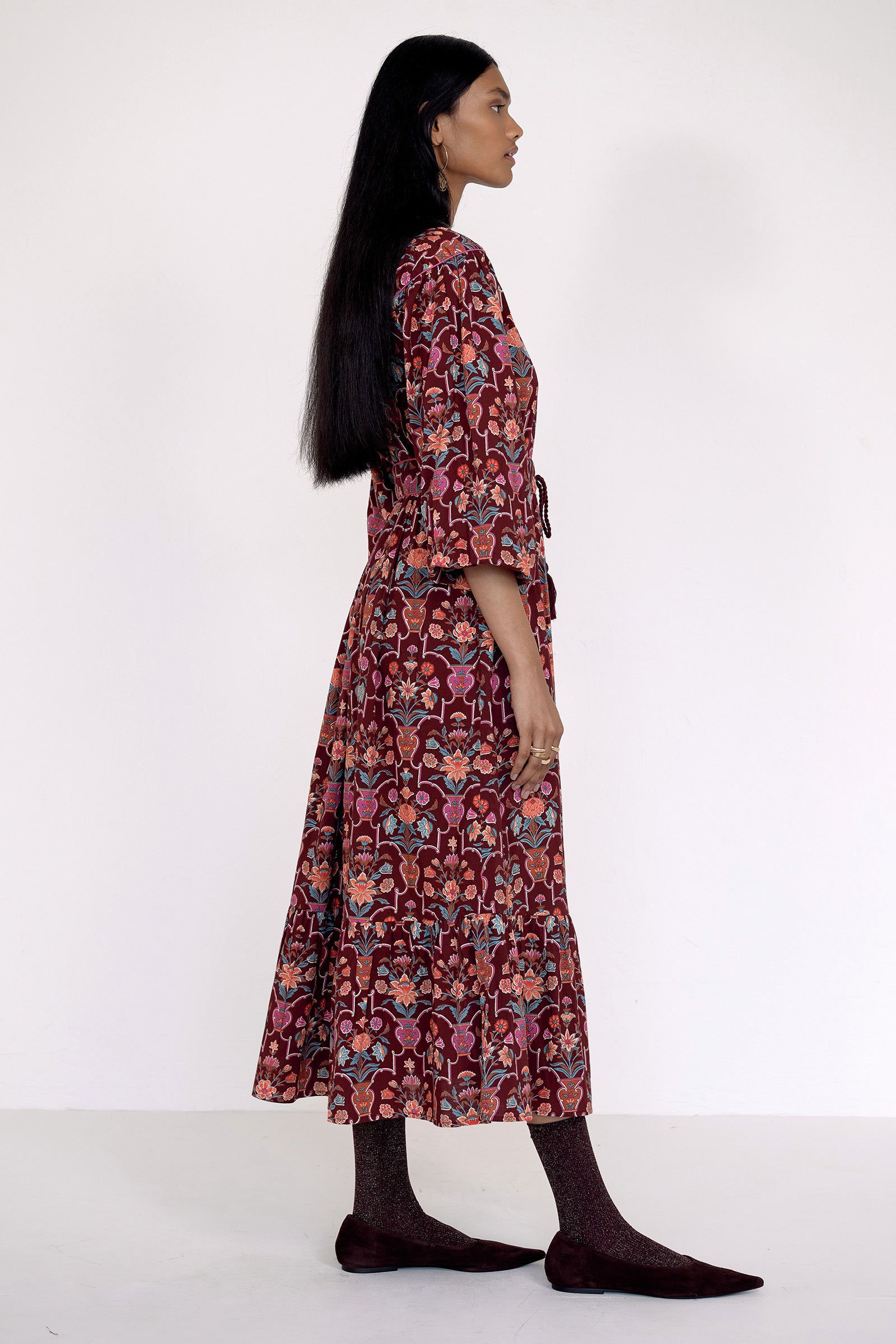 Side view of model standing wearing the Victoria dress in Floral Vase print. It features quilted shoulder details, piping, a tassel waist tie, and an easy silhouette.