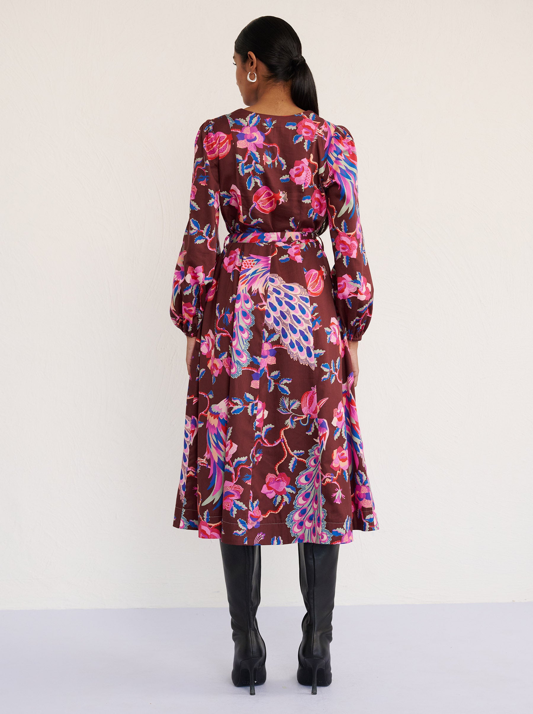 Back view of a model standing. She is wearing the Zora dress in the Bukhara Peacock print in rum color. It is featuring lantern sleeves, a V-neckline, princess seams, a fit-and-flare skirt, a fabric belt, and side-seam pockets.