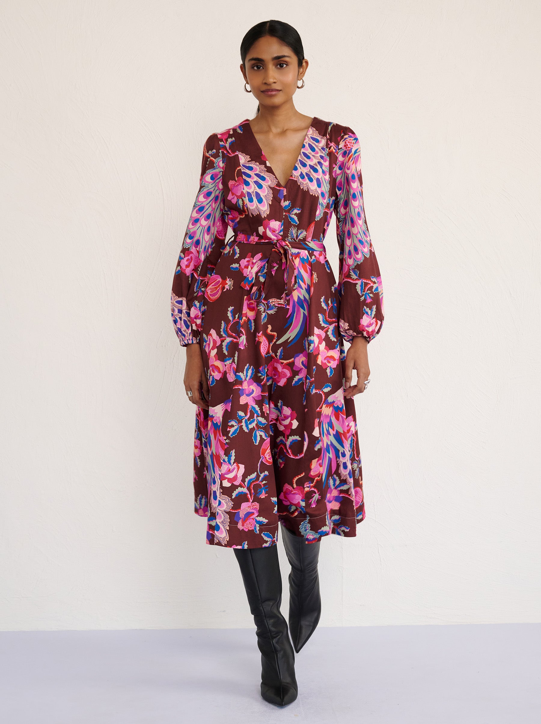 Front view of a model walking. She is wearing the Zora dress in the Bukhara Peacock print in rum color. It is featuring lantern sleeves, a V-neckline, princess seams, a fit-and-flare skirt, a fabric belt, and side-seam pockets.