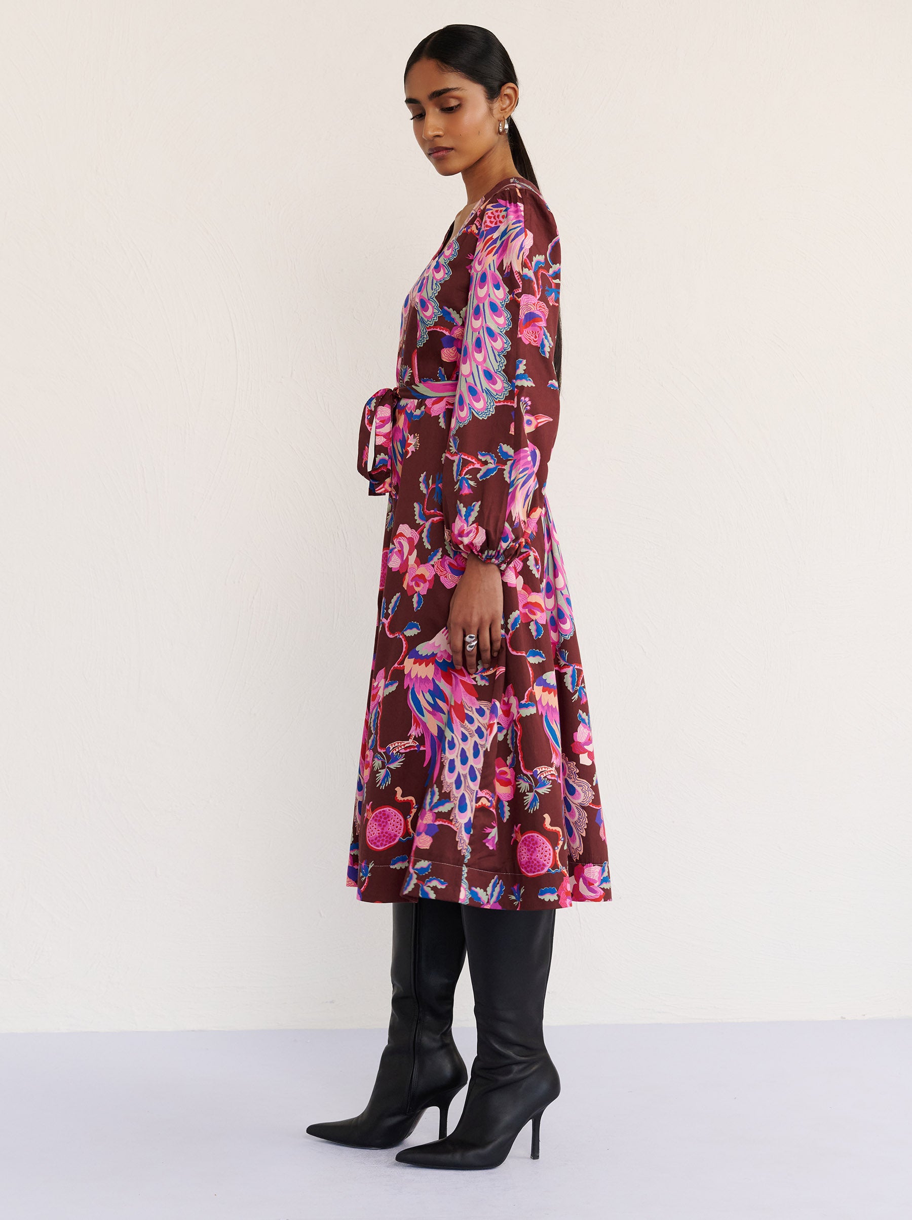 Side view of a model standing. She is wearing the Zora dress in the Bukhara Peacock print in rum color. It is featuring lantern sleeves, a V-neckline, princess seams, a fit-and-flare skirt, a fabric belt, and side-seam pockets.