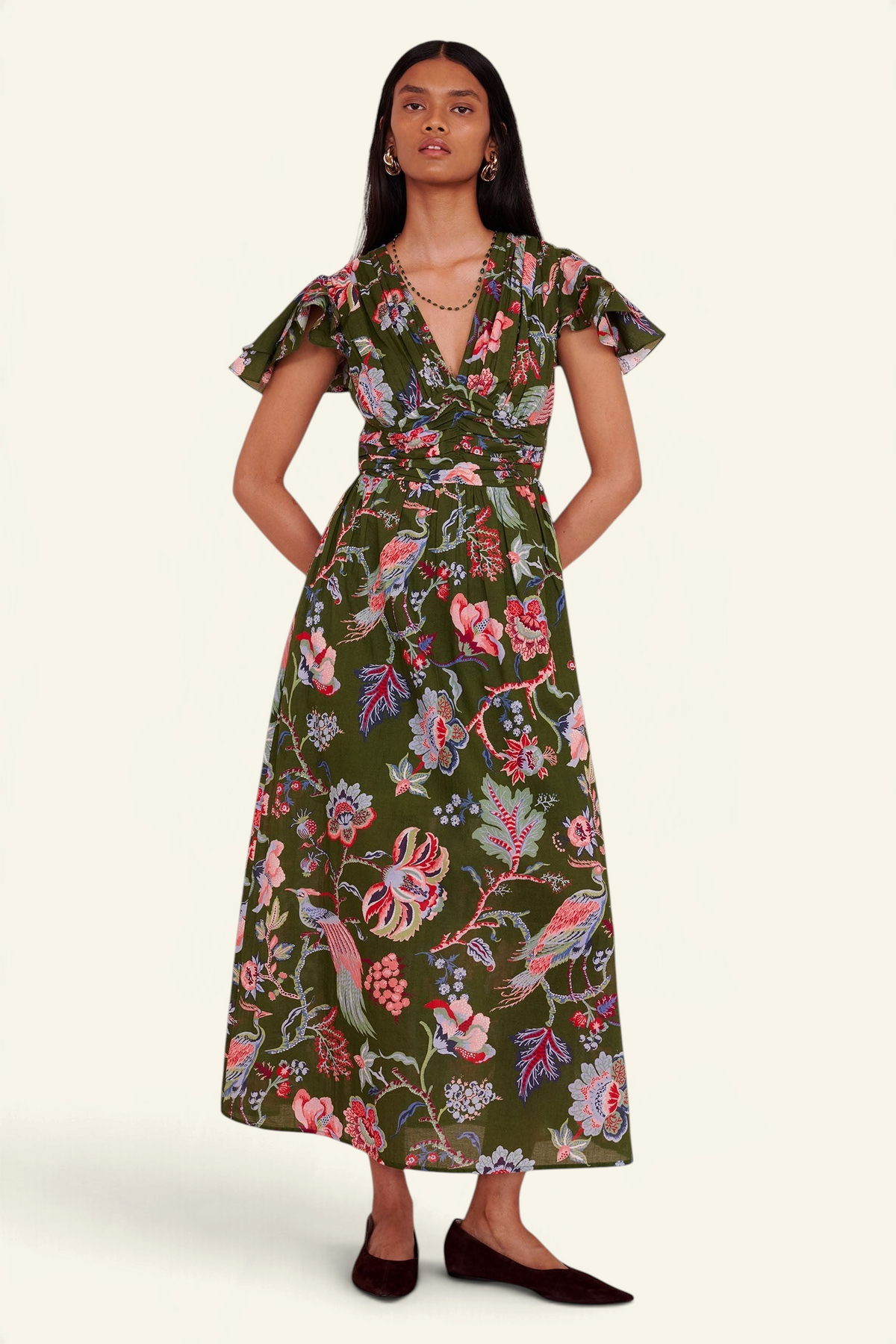 A woman modeling a dark olive green midi dress with a vibrant pink, red, and blue floral print. The dress features short flutter sleeves, a V-neckline, and a self-tie waist.