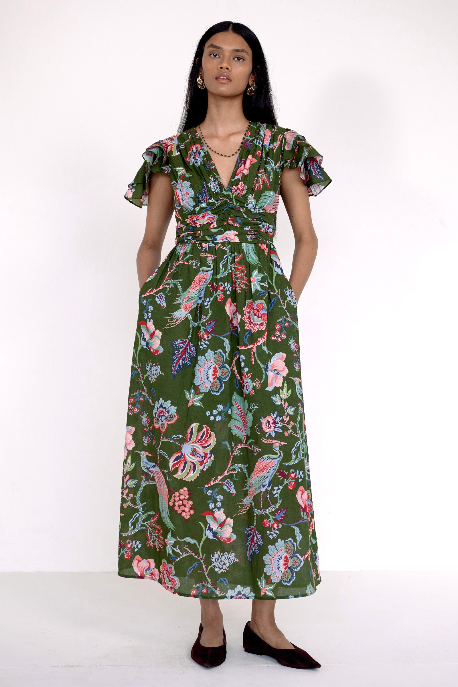 Front view of model standing. She is  wearing the Adele dress in Chintz print moss color with hands in the pocket. It features a V-neck, pleat details across the bust, and delicate flutter sleeves.