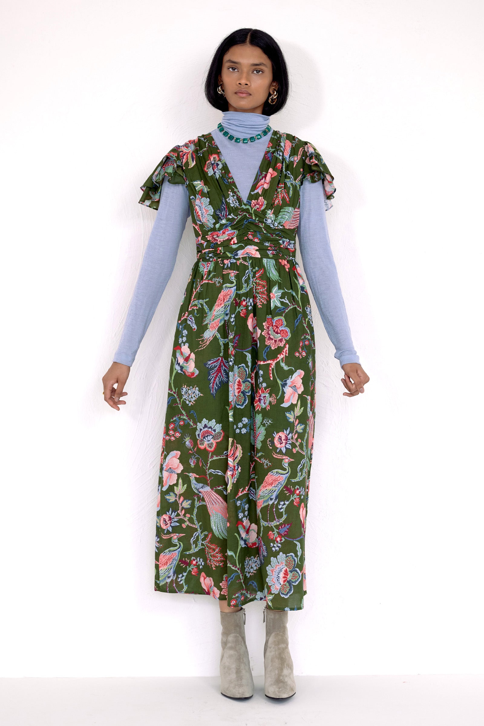 Front view of model standing. She is  wearing the Adele dress in Chintz print moss color. It features a V-neck, pleat details across the bust, and delicate flutter sleeves. Its styled with a nigh neck layer inside with boots and accessories.