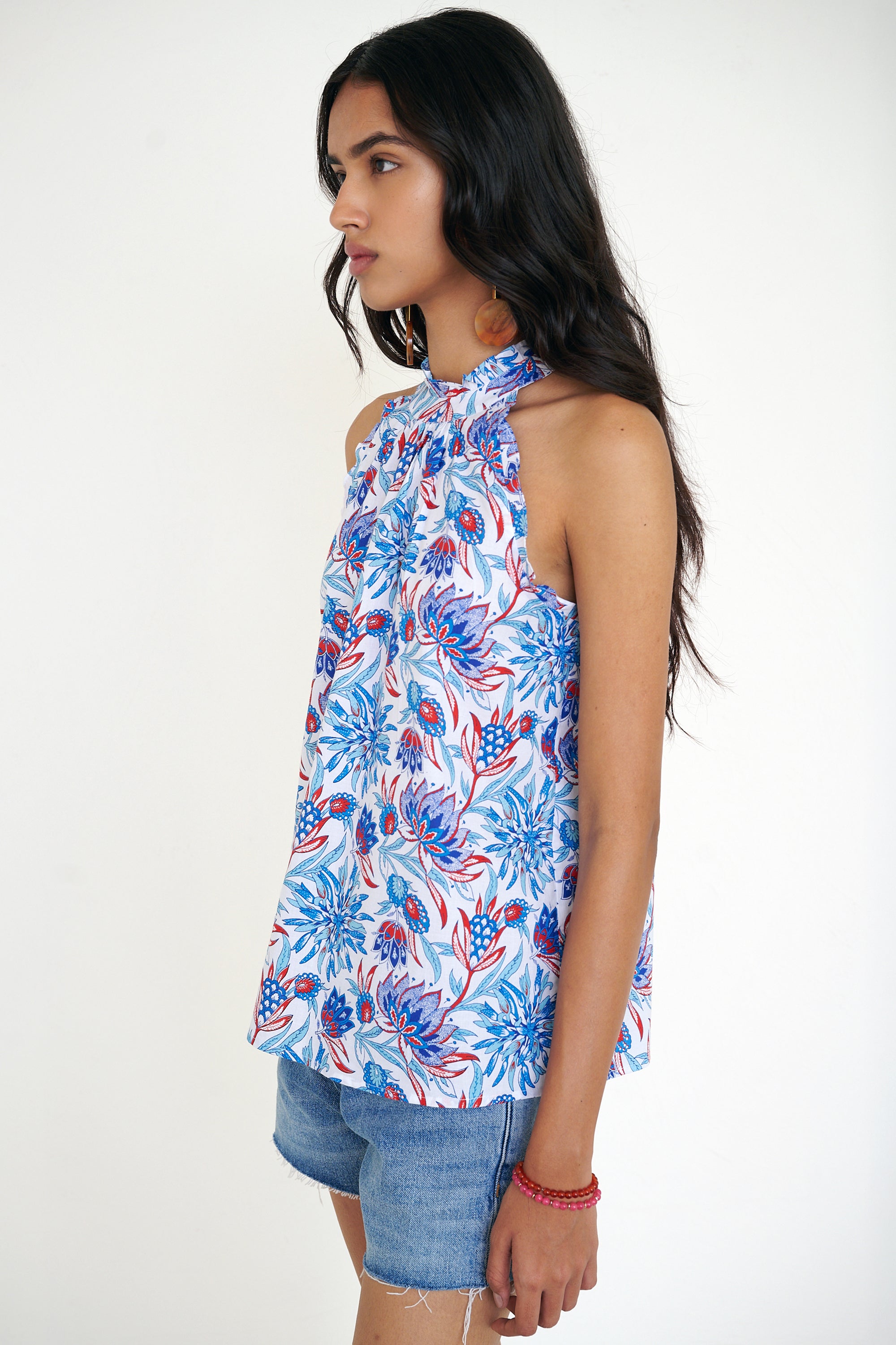 Side view of woman standing wearing the Amy top in the Philippa print, featuring a sleeveless silhouette, subtle ruffle neckline, and a red, white, and blue wildflower pattern. Paired with denim shorts.