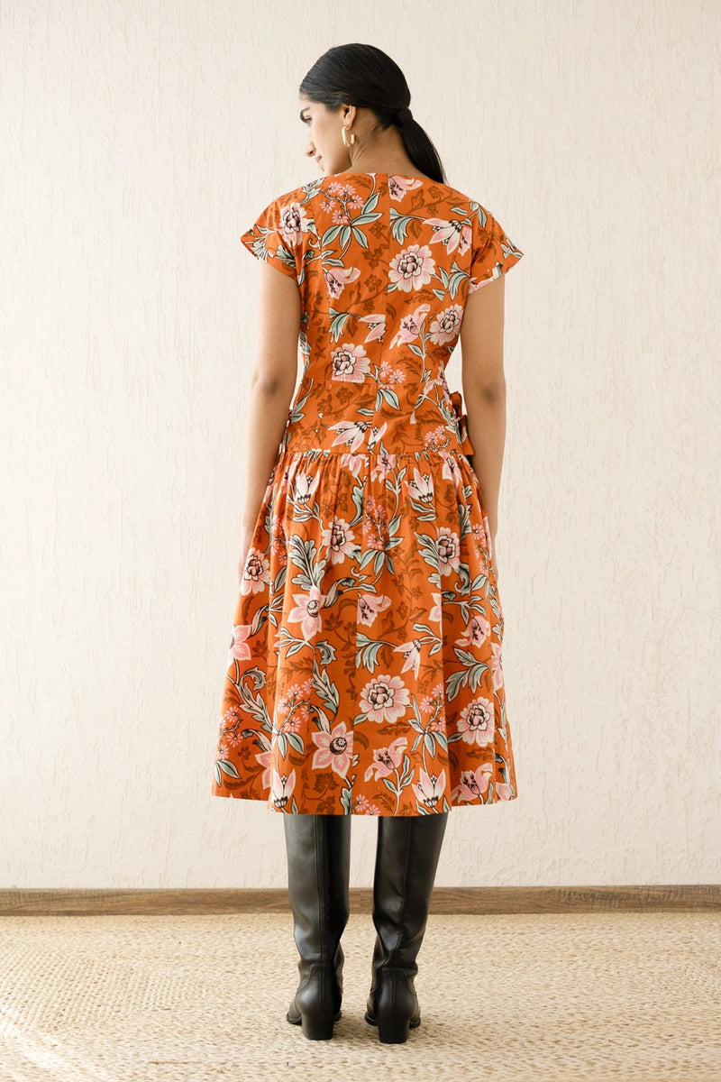 Back view of model wearing the Angela dress with a V-neck, drop waist, side ties, and bold floral print, made from cotton poplin. Perfect for layering in autumn.