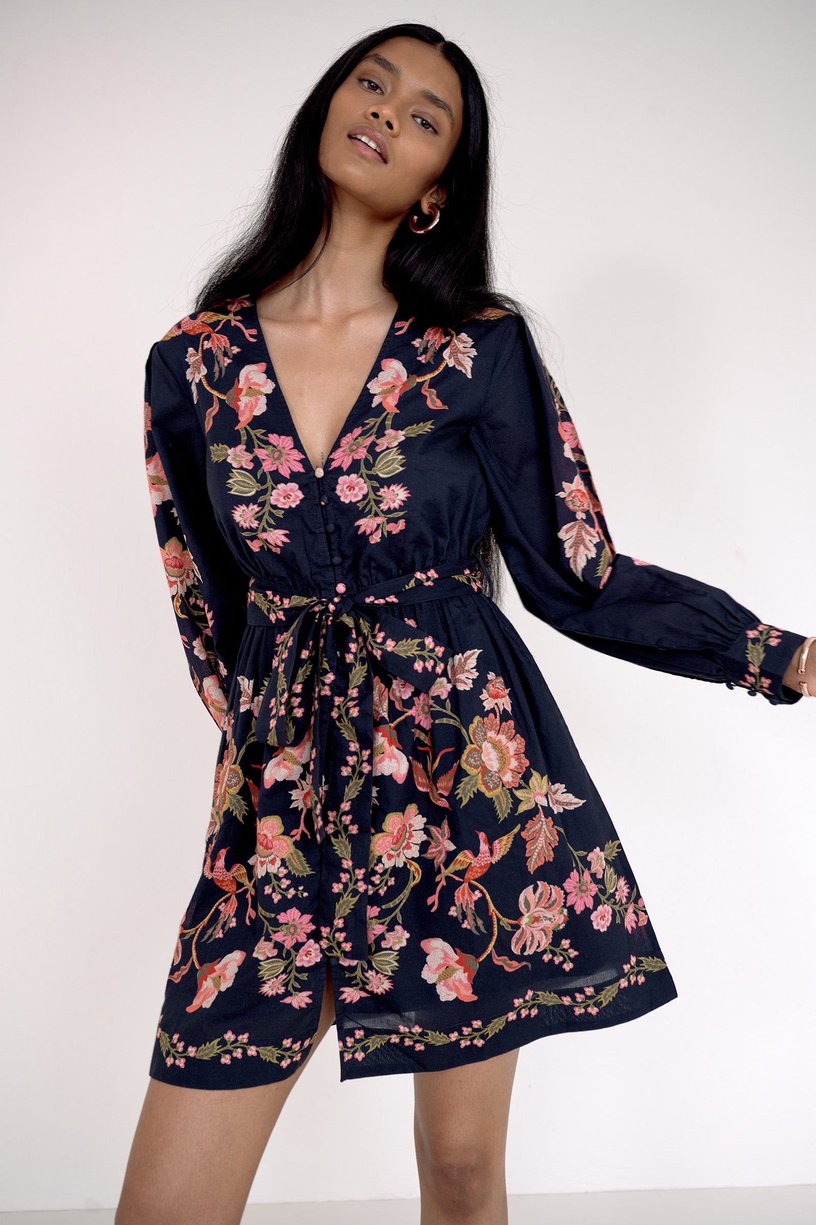 Front view close up of model. She is wearing the Anne dress in Chintz print. It features long sleeves, an elasticated waist, fabric-covered buttons, a fabric belt, and side-seam pockets.