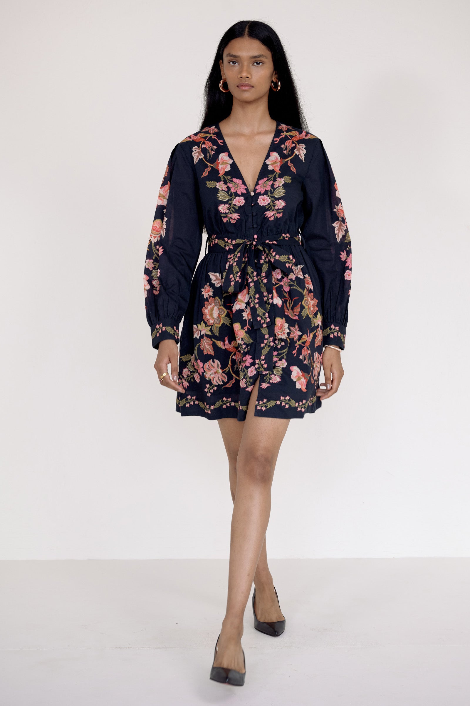 Front view of model walking forward. She is wearing the Anne dress in Chintz print. It features long sleeves, an elasticated waist, fabric-covered buttons, a fabric belt, and side-seam pockets.