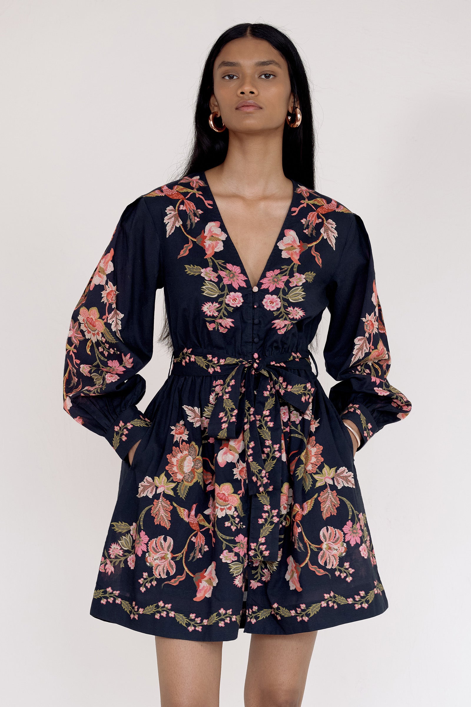 Front view close up of model standing. She is wearing the Anne dress in Chintz print with hands in pocket. It features long sleeves, an elasticated waist, fabric-covered buttons, a fabric belt, and side-seam pockets.