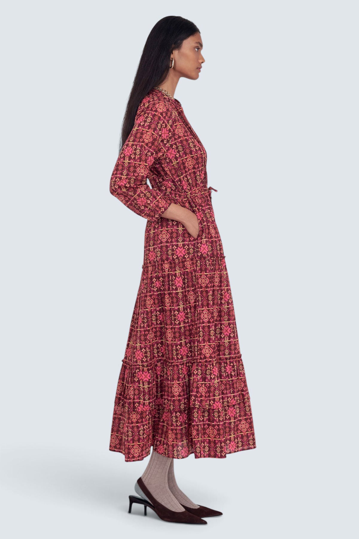 Back view of model standing wearing the Bazaar dress in Needlepoint print, featuring long sleeves, a tiered ankle-length A-line skirt, drawstring belt, and side-seam pockets.