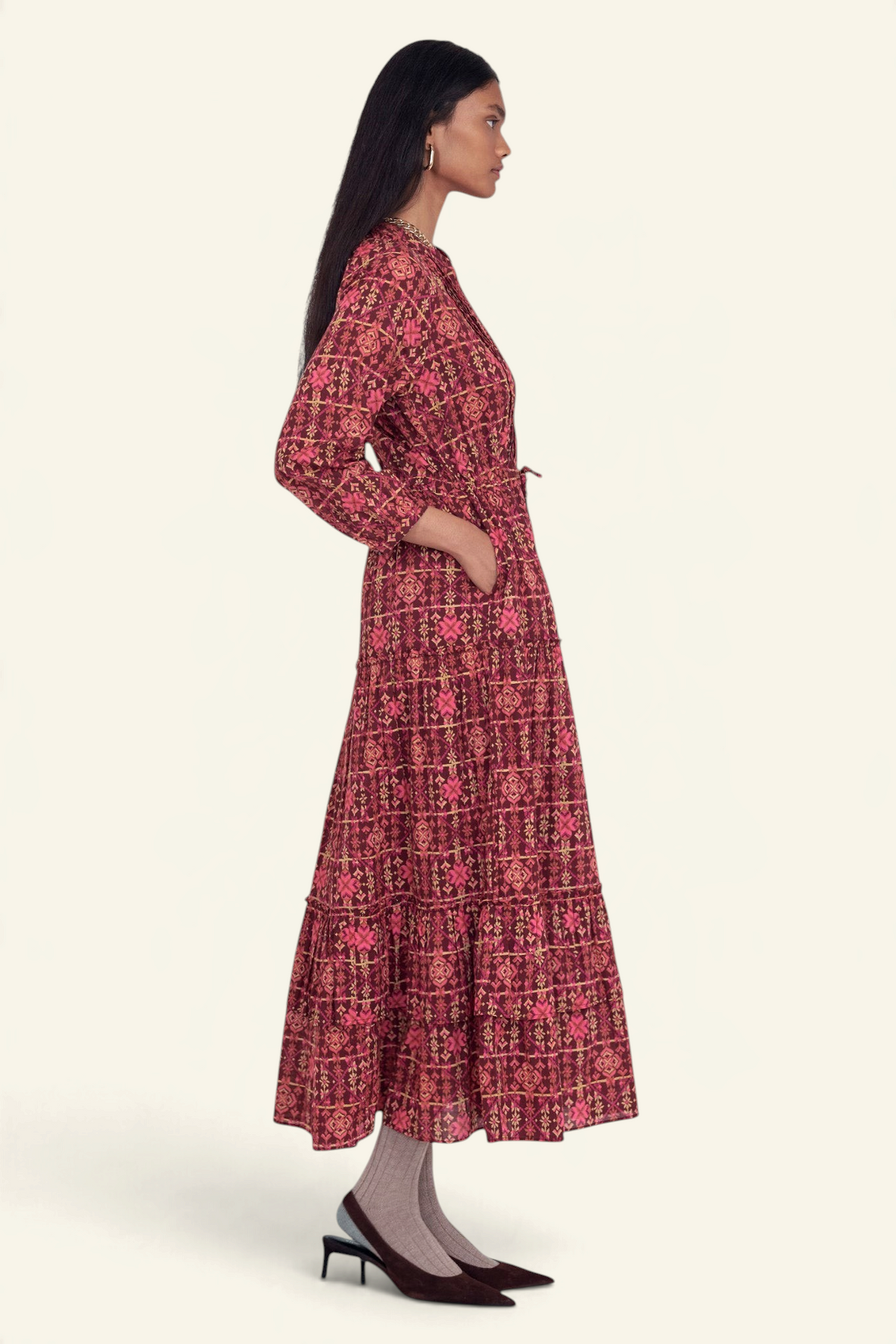 Back view of model standing wearing the Bazaar dress in Needlepoint print, featuring long sleeves, a tiered ankle-length A-line skirt, drawstring belt, and side-seam pockets.