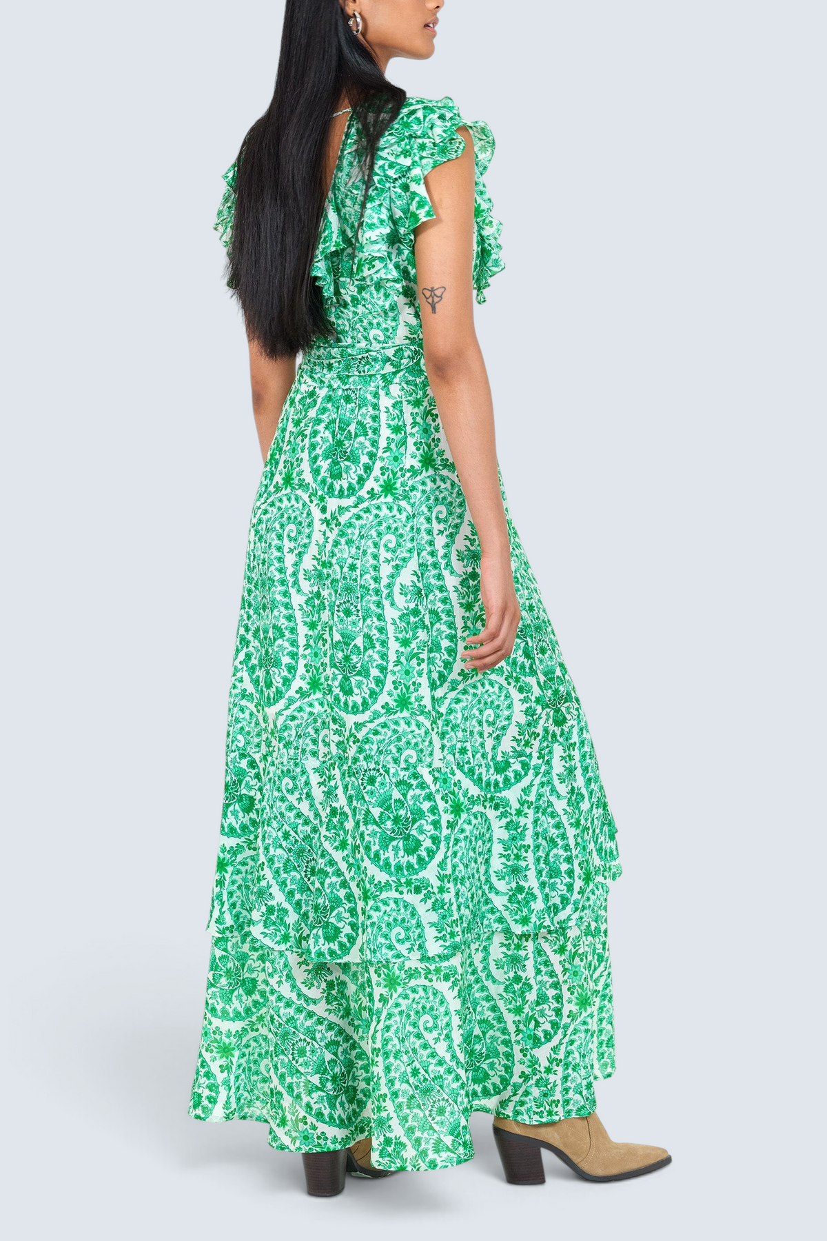 Side view of a woman standing. She is  wearing the Beatrice dress in Paisley print, featuring a full-length tiered skirt, ruffled bodice, fabric tie belt, dramatic tie-back neckline, and lustrous cotton silk.