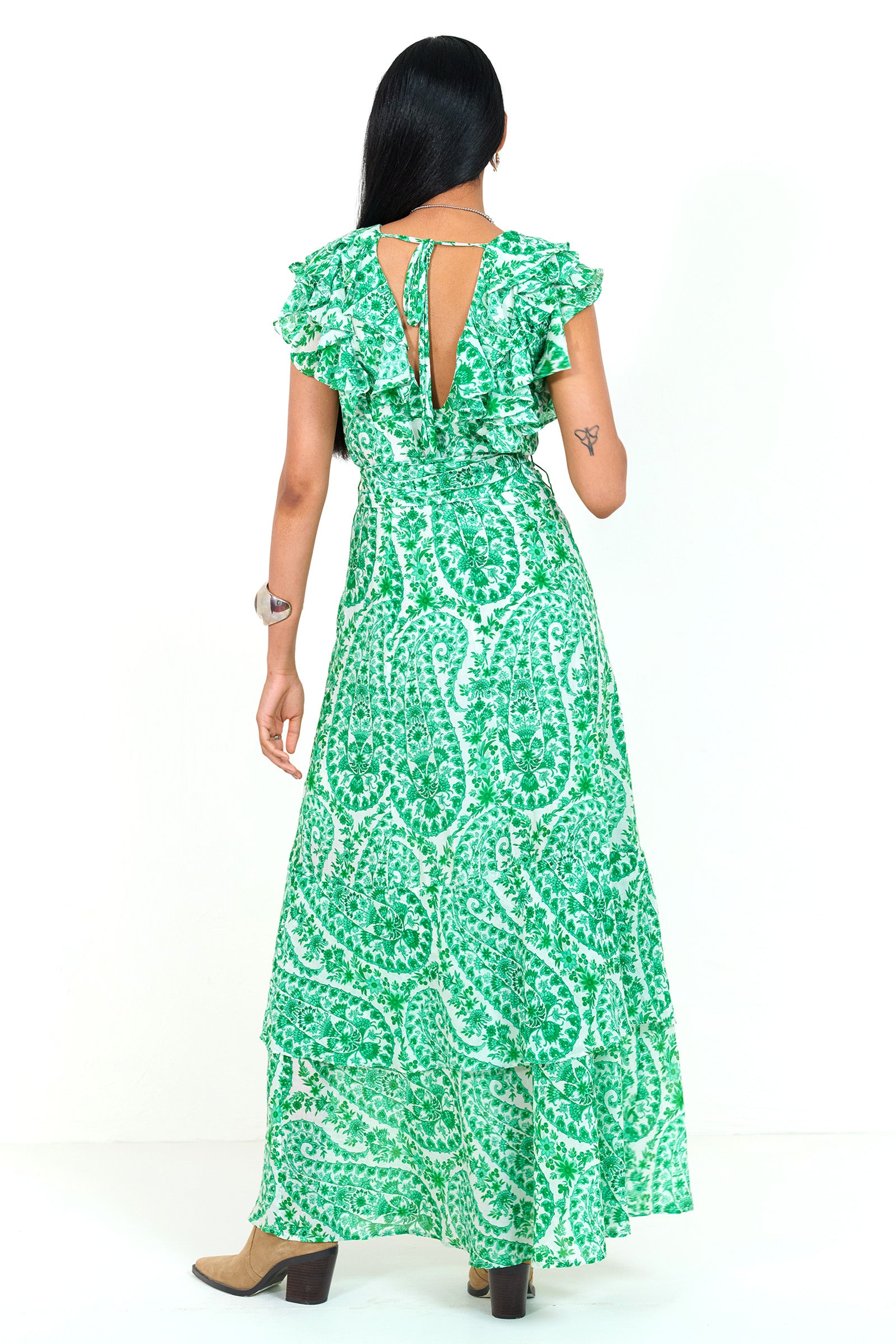 Back view of a woman standing. She is  wearing the Beatrice dress in Paisley print, featuring a full-length tiered skirt, ruffled bodice, fabric tie belt, dramatic tie-back neckline, and lustrous cotton silk.