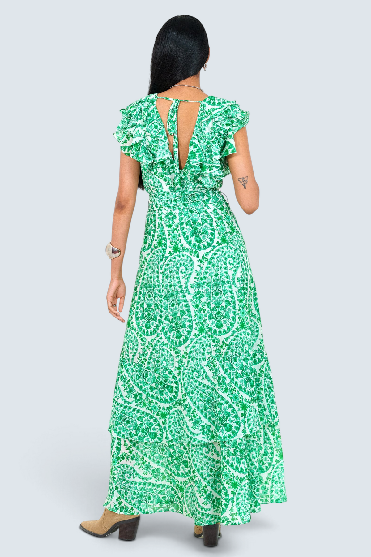 Back view of a woman standing. She is  wearing the Beatrice dress in Paisley print, featuring a full-length tiered skirt, ruffled bodice, fabric tie belt, dramatic tie-back neckline, and lustrous cotton silk.