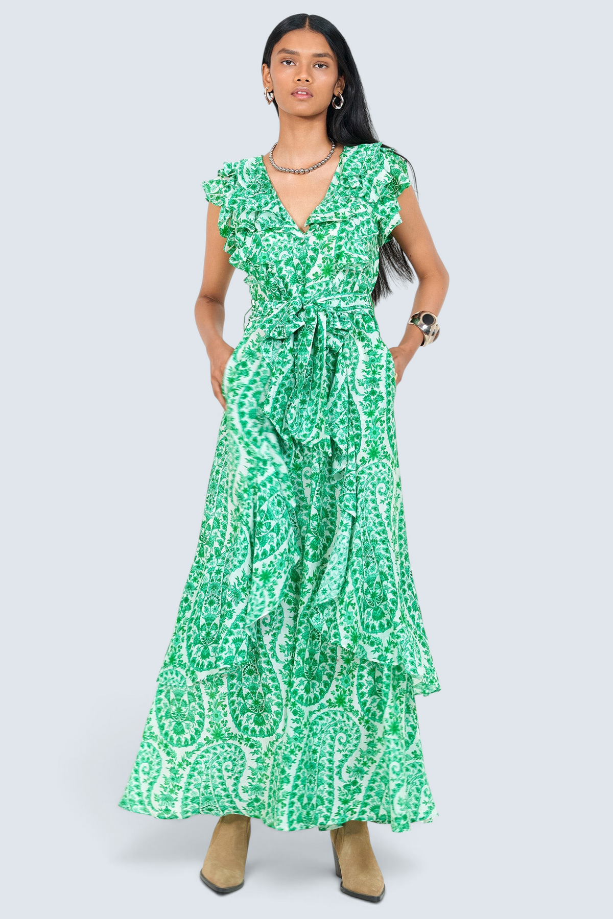 Front view of a woman standing. She is  wearing the Beatrice dress in Paisley print with hands in pocket. It features a full-length tiered skirt, ruffled bodice, fabric tie belt, dramatic tie-back neckline, and lustrous cotton silk.