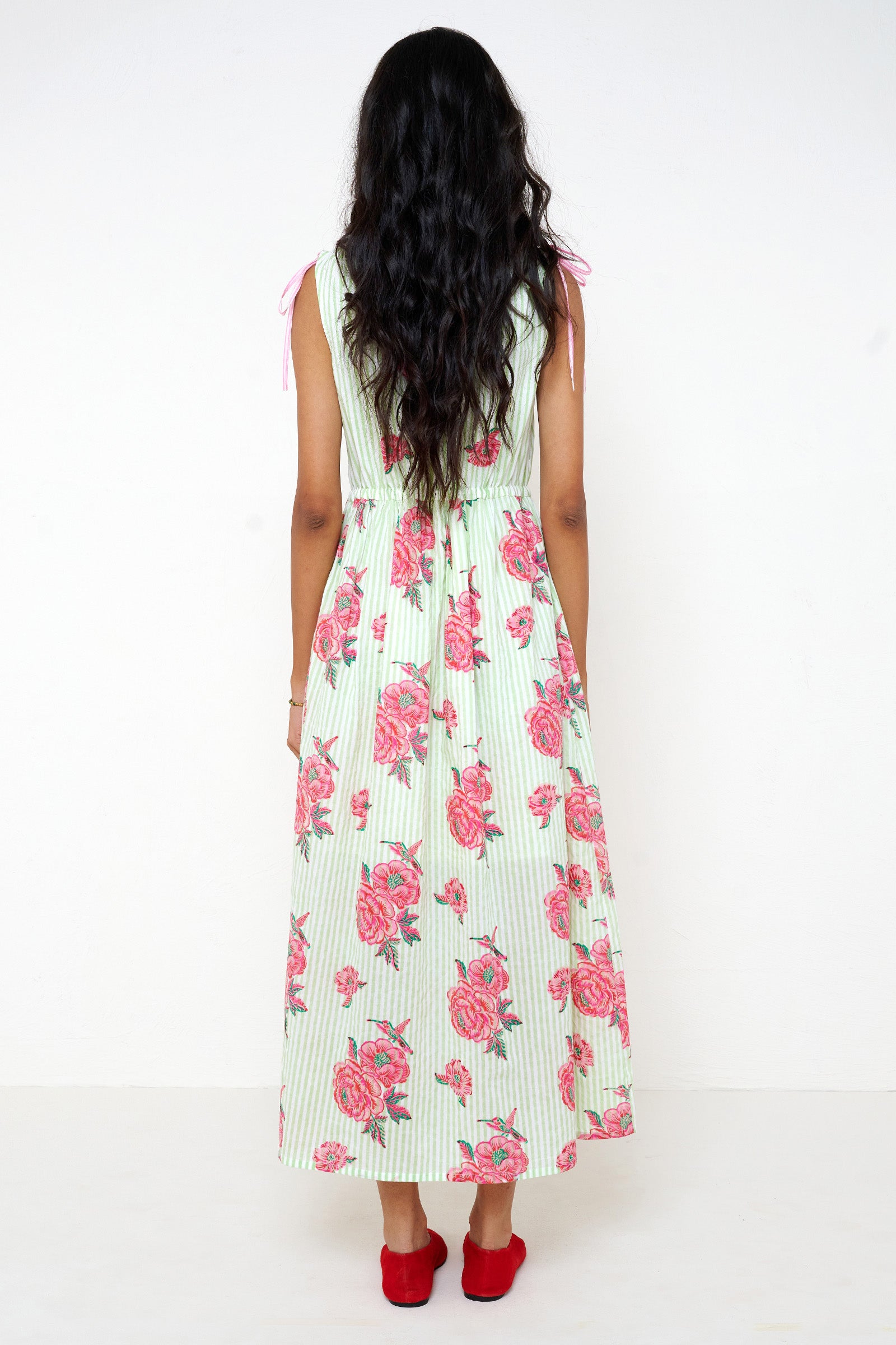Back view of woman standing. She is wearing the Becca dress in the Eliza Rose print, featuring an adjustable waist, shoulder ties, a deep V-neckline, and hand block-printed floral motifs with hummingbird details.