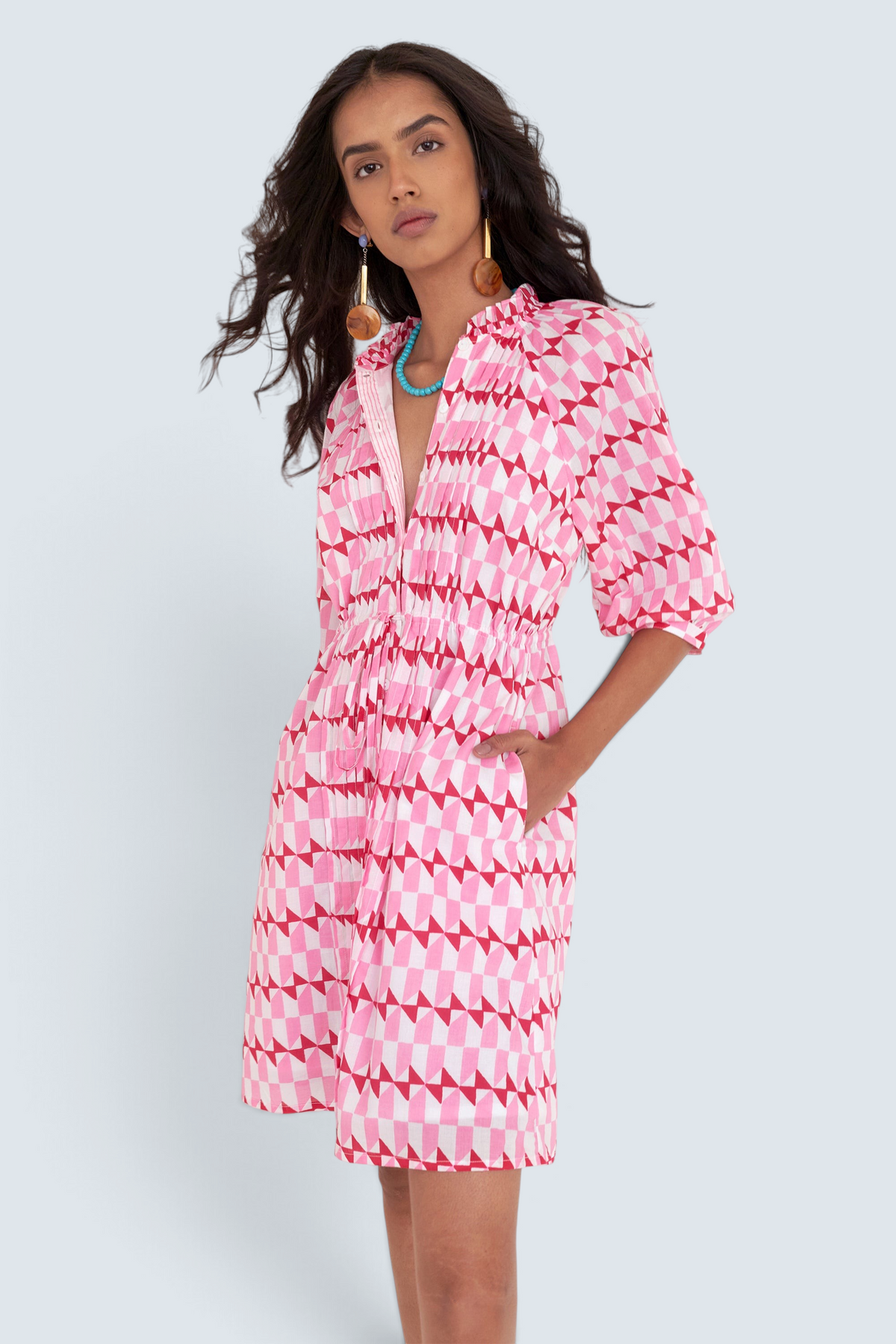 Front side view of woman standing. She is  wearing the Benita dress in the Stevie print, featuring a shirtdress silhouette, button-front placket, drawstring waist, and geometric motifs inspired by the French Riviera.