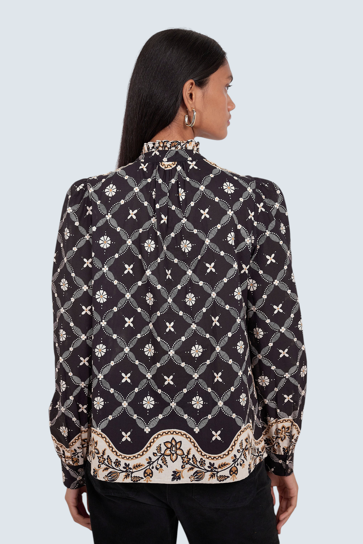 Back view of model standing. She is wearing Banjanan Christina shirt in black with a Victorian-inspired geometric bandana print, frilled collar, and puff sleeves.