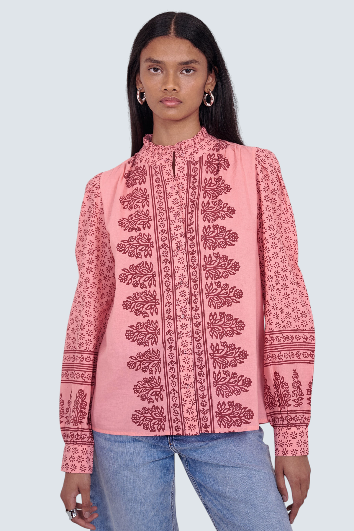Front view close up of model standing. She is wearing the Christina shirt, featuring a frilled stand collar, gathered neckline, slightly puffed sleeves, and hand-placed geometric floral block print. Its paired with jeans.