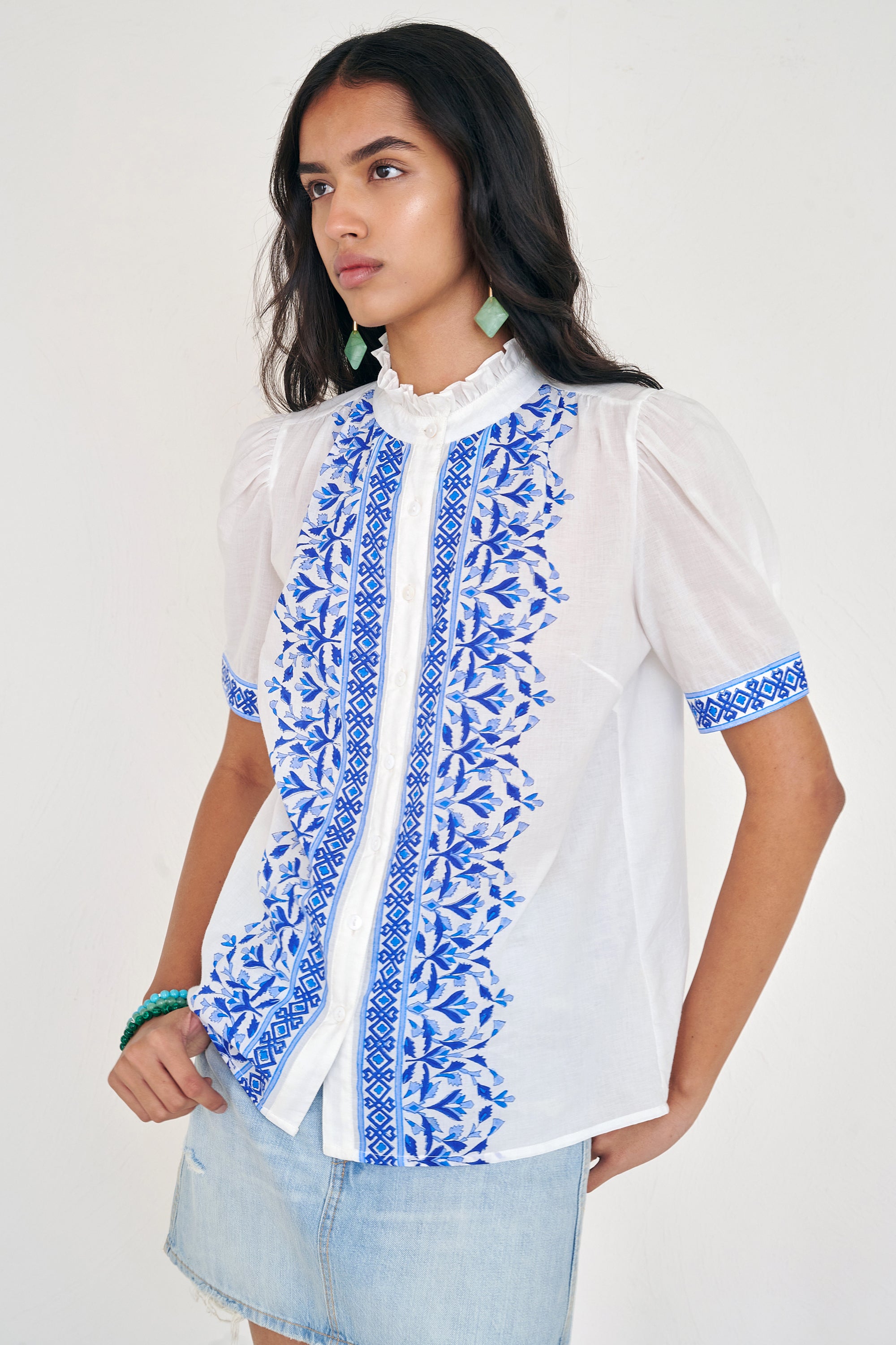 Front side view of woman standing. She is  wearing the Christina shirt, featuring a hand block-printed geometric floral pattern, a frilled stand collar, soft gathers at the neckline, and slightly puffed sleeves. Paired with denim skirt.