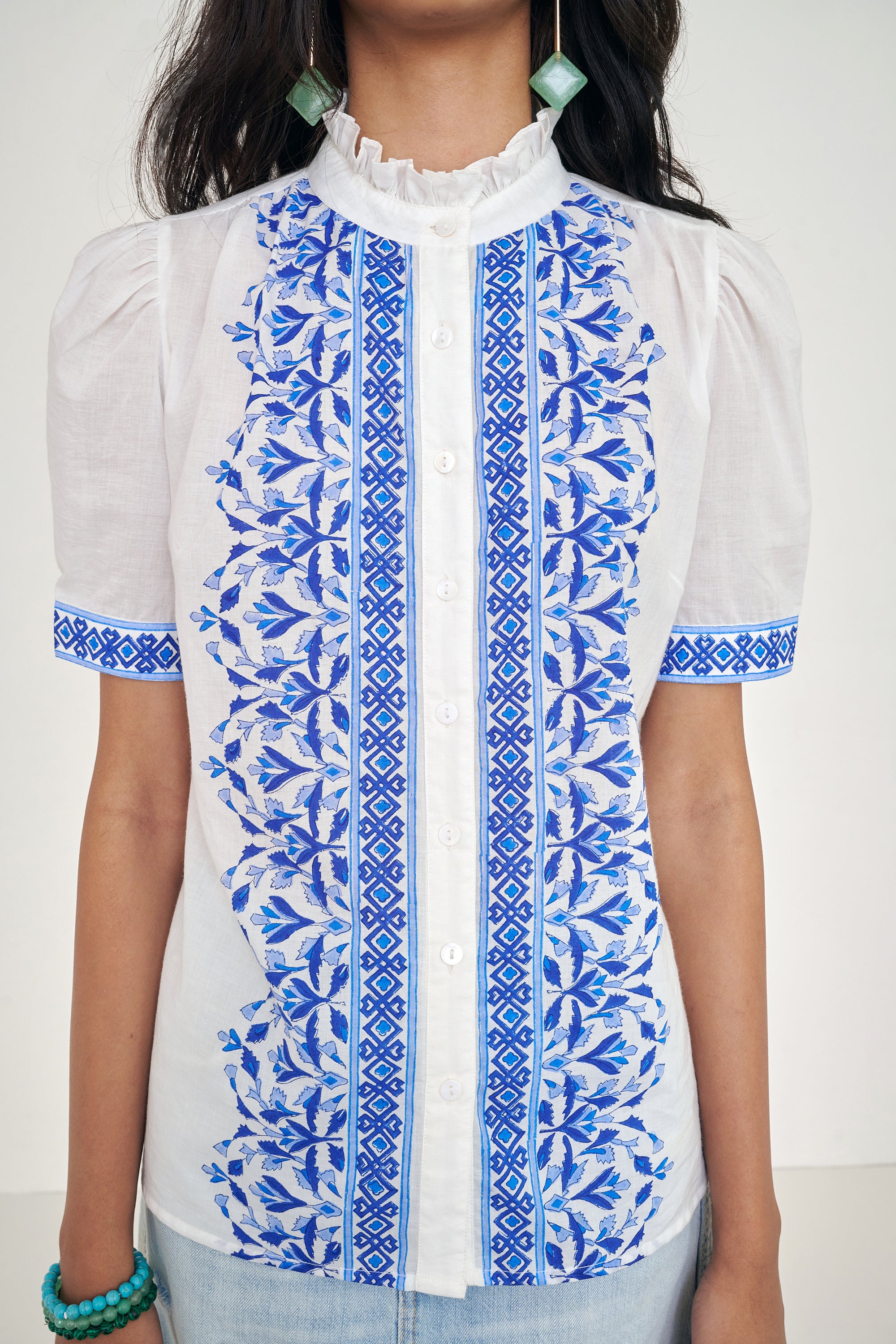 Close up of front view of woman standing. She is  wearing the Christina shirt, featuring a hand block-printed geometric floral pattern, a frilled stand collar, soft gathers at the neckline, and slightly puffed sleeves. Paired with denim skirt.