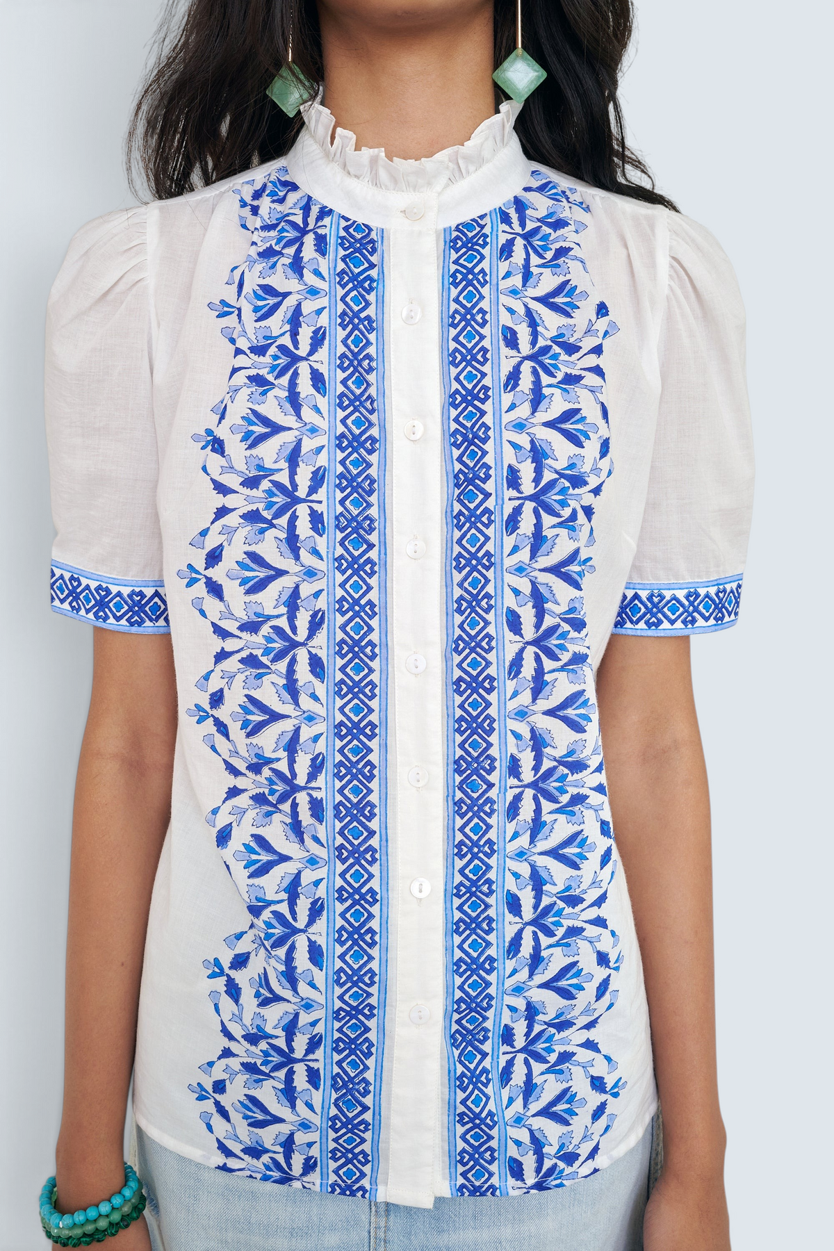 Close up of front view of woman standing. She is  wearing the Christina shirt, featuring a hand block-printed geometric floral pattern, a frilled stand collar, soft gathers at the neckline, and slightly puffed sleeves. Paired with denim skirt.