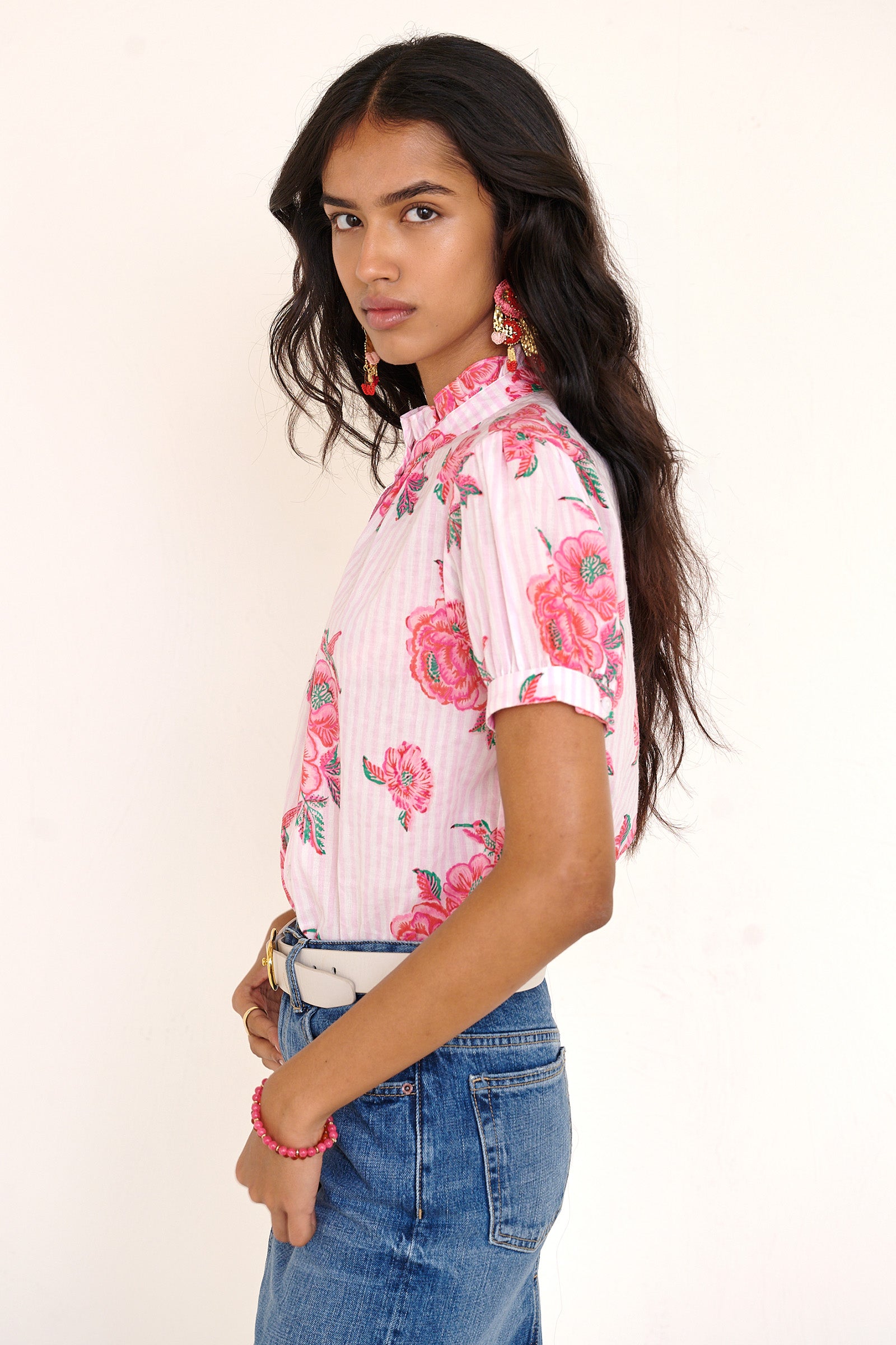 Side view of woman standing wearing the Christina shirt, featuring hand block-printed rose motifs, a ruffled neckline, subtly puffed short sleeves, and a button-front silhouette.