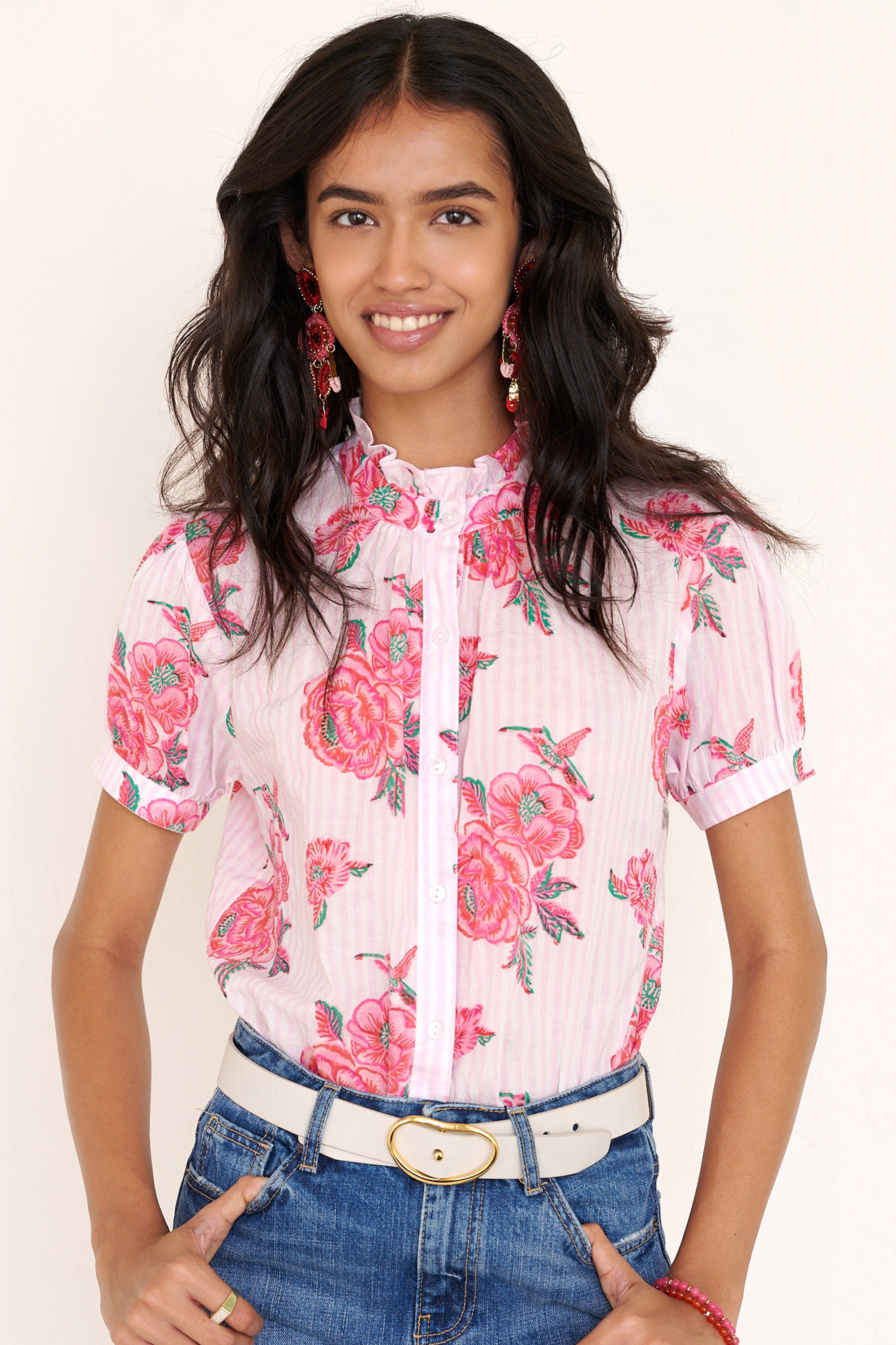 Front view of woman standing wearing the Christina shirt, featuring hand block-printed rose motifs, a ruffled neckline, subtly puffed short sleeves, and a button-front silhouette.