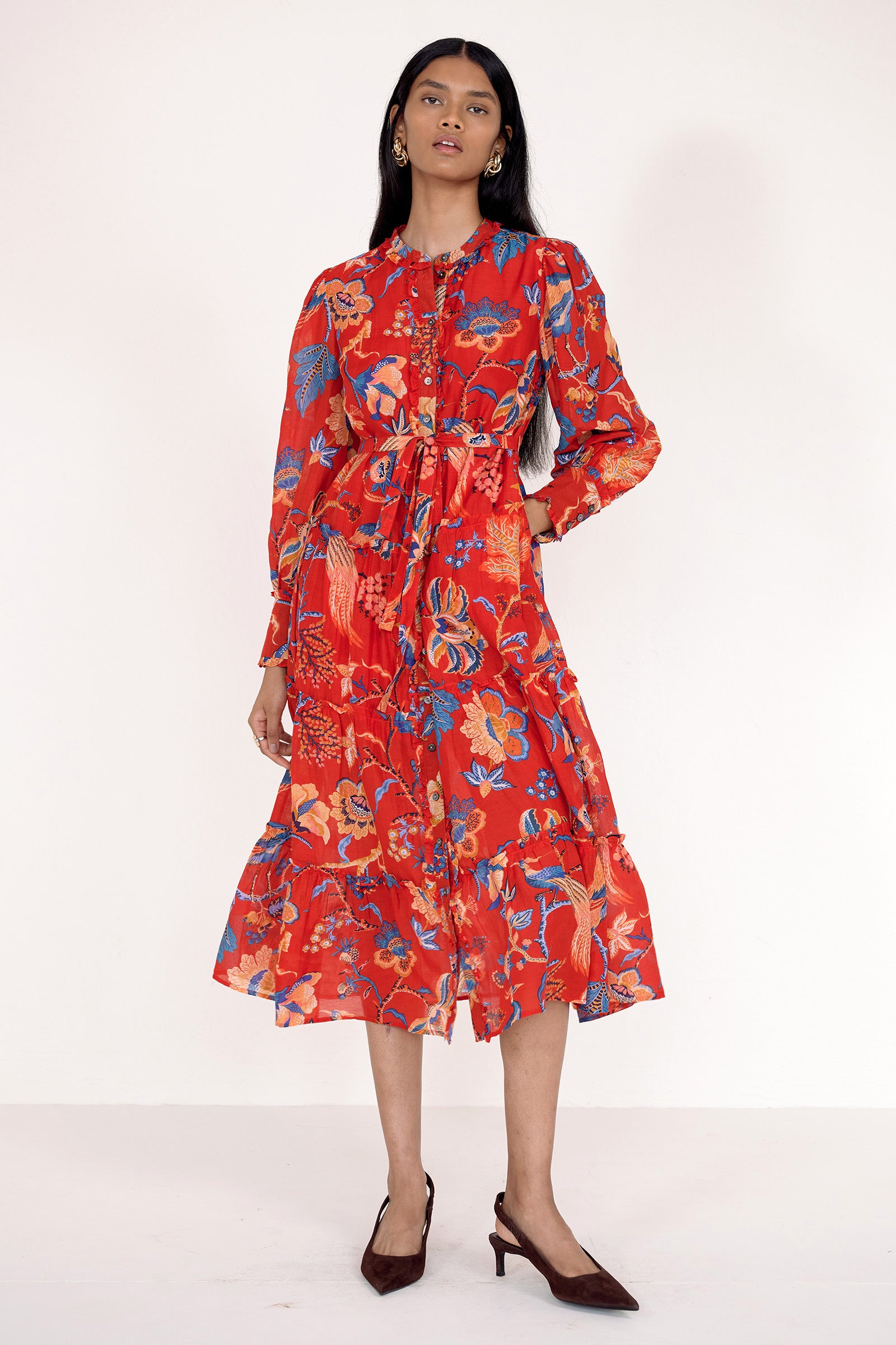 Front view of model standing. She is wearing the Emira dress in Chintz print with left hand in pocket. It features a flowing skirt, delicate ruffles, fabric-covered buttons, and a detachable belt.