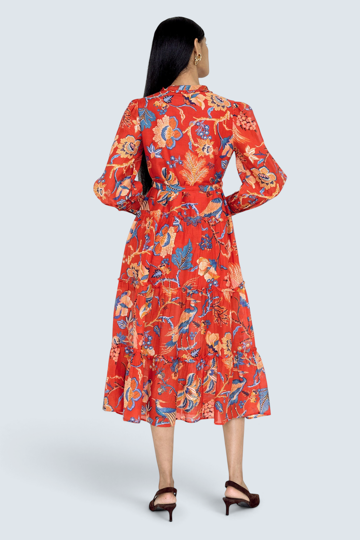 Back view of model standing. She is wearing the Emira dress in Chintz print. It  features a flowing skirt, delicate ruffles, fabric-covered buttons, and a detachable belt.