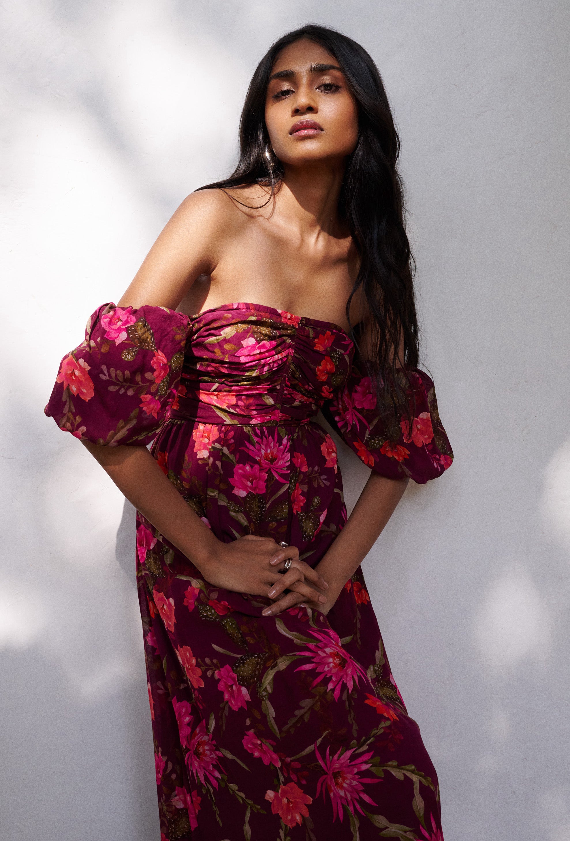 A model is standing in front of a white wall posing wearing Farida Dress in Desert Rose print. It has a nice off shoulder detail and a ruched bodice.
