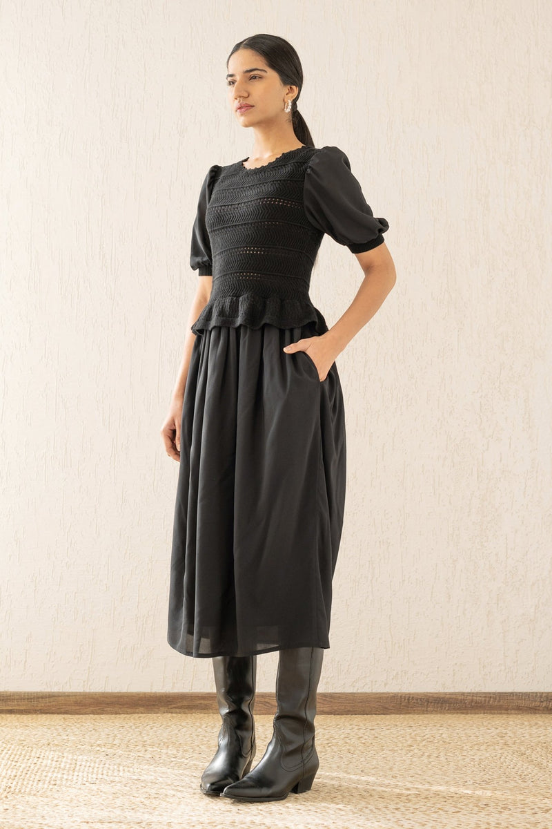 Side view of a woman wearing the Heather maxi dress in black, featuring hand-crocheted details, a woven viscose georgette skirt, and a soft peplum waist for a flattering silhouette.