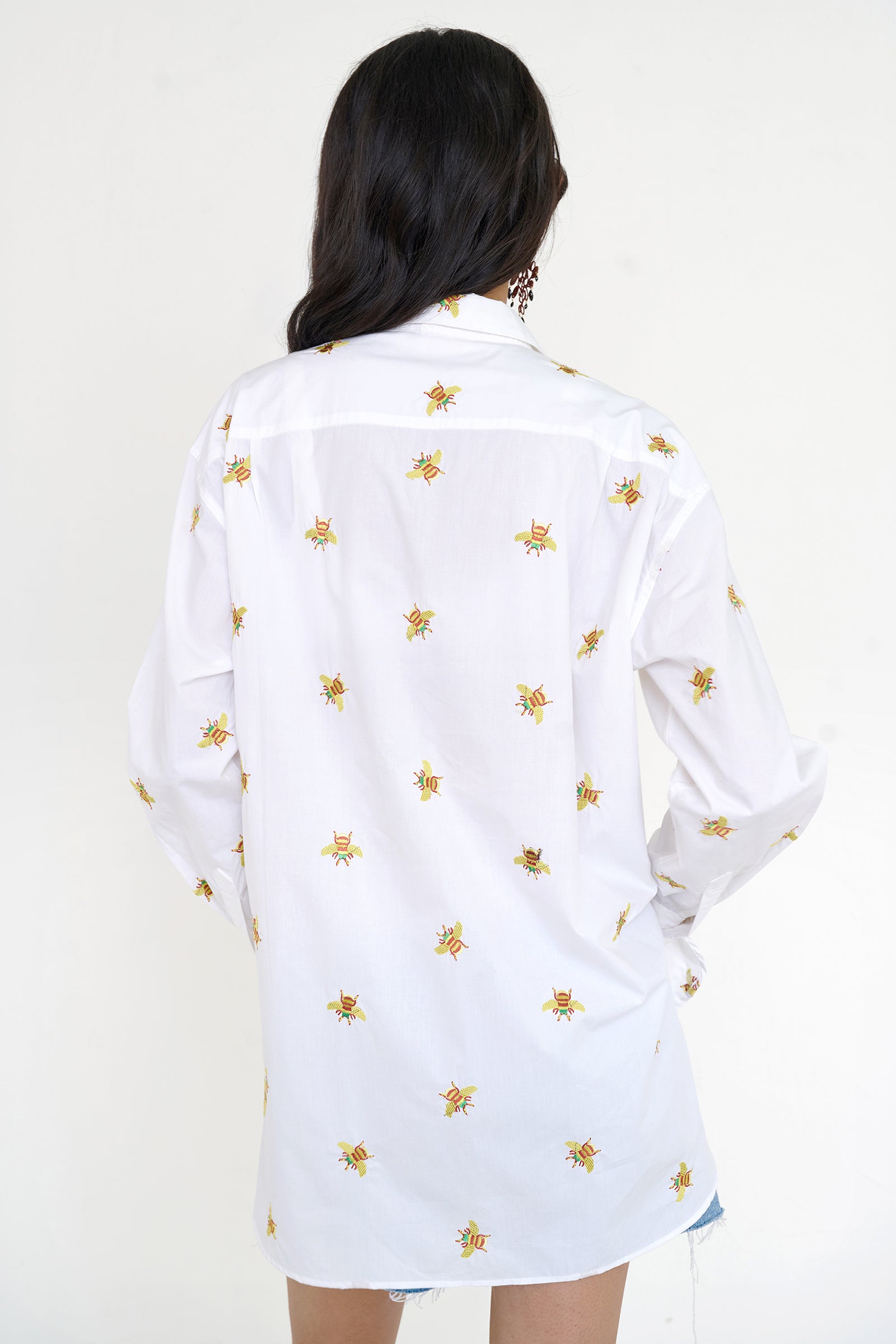 Back view of woman standing wearing the Jamie shirt, featuring a menswear-inspired button-up silhouette, embroidered bumblebee motifs, pleat details, and a high-low hem.